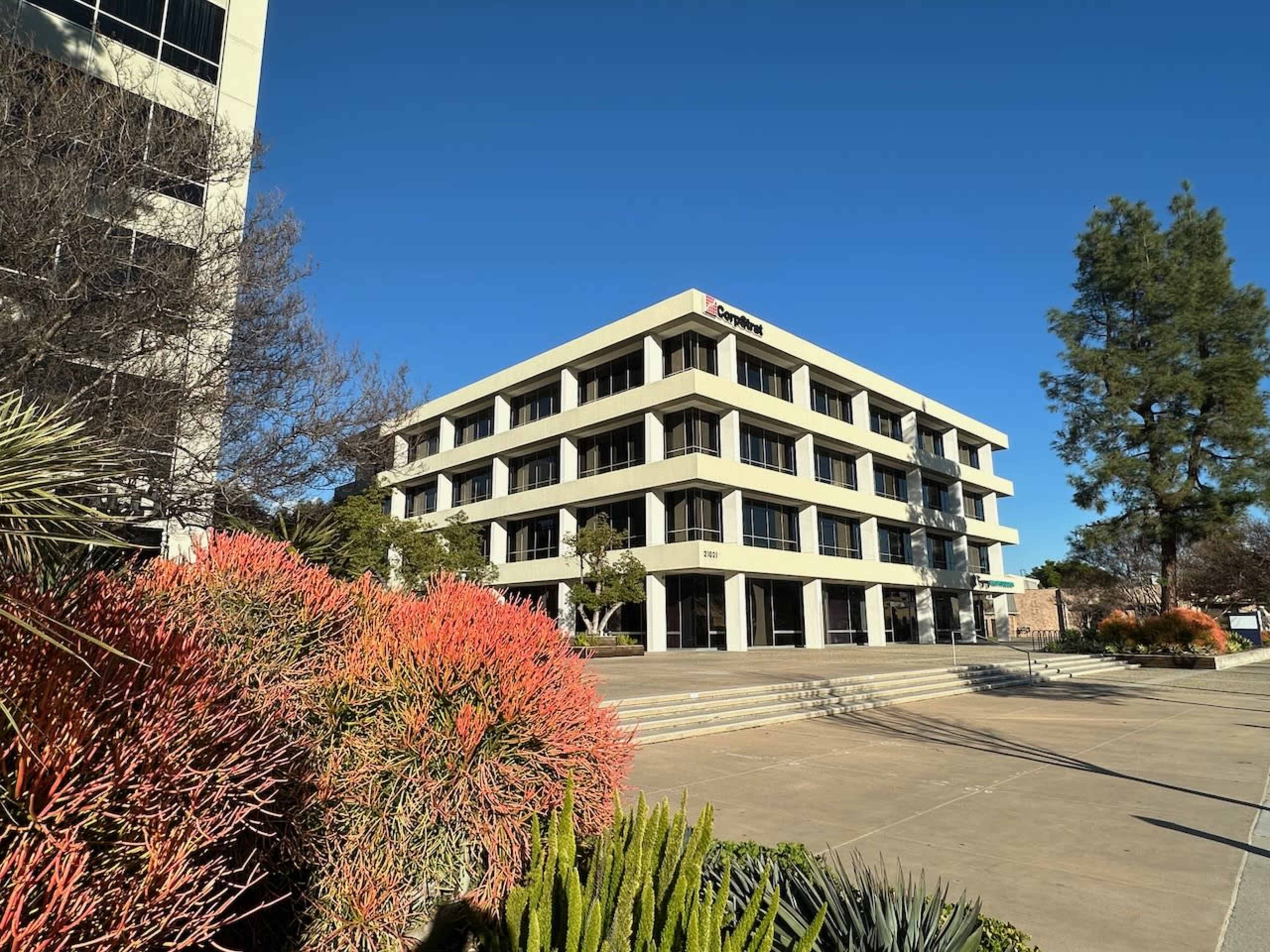 A modern, four-story office building is set against a clear blue sky, surrounded by landscaped greenery and colorful plants.