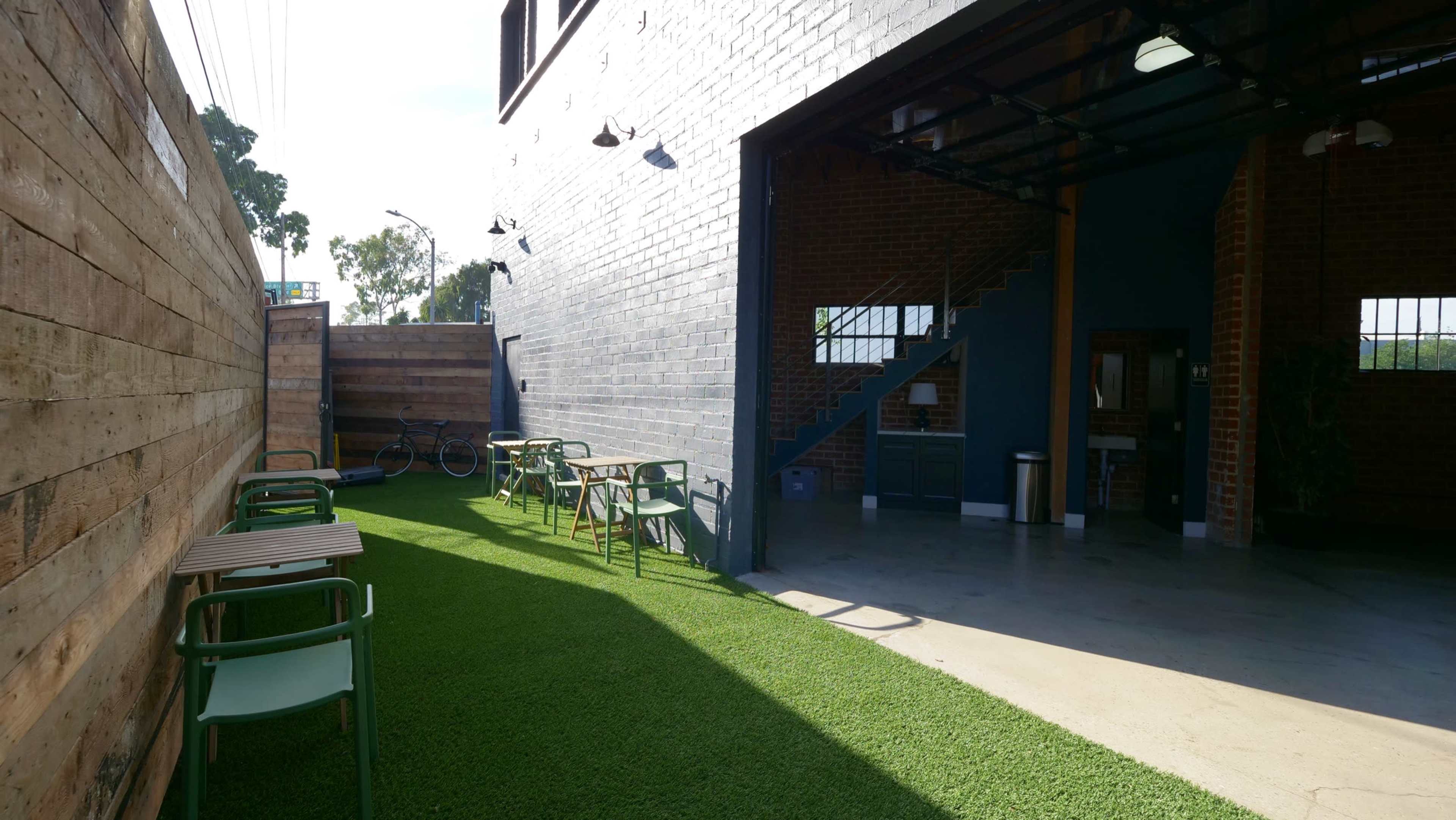 The image shows an outdoor seating area with green chairs and wooden tables adjacent to a modern building with a partially open garage door.
