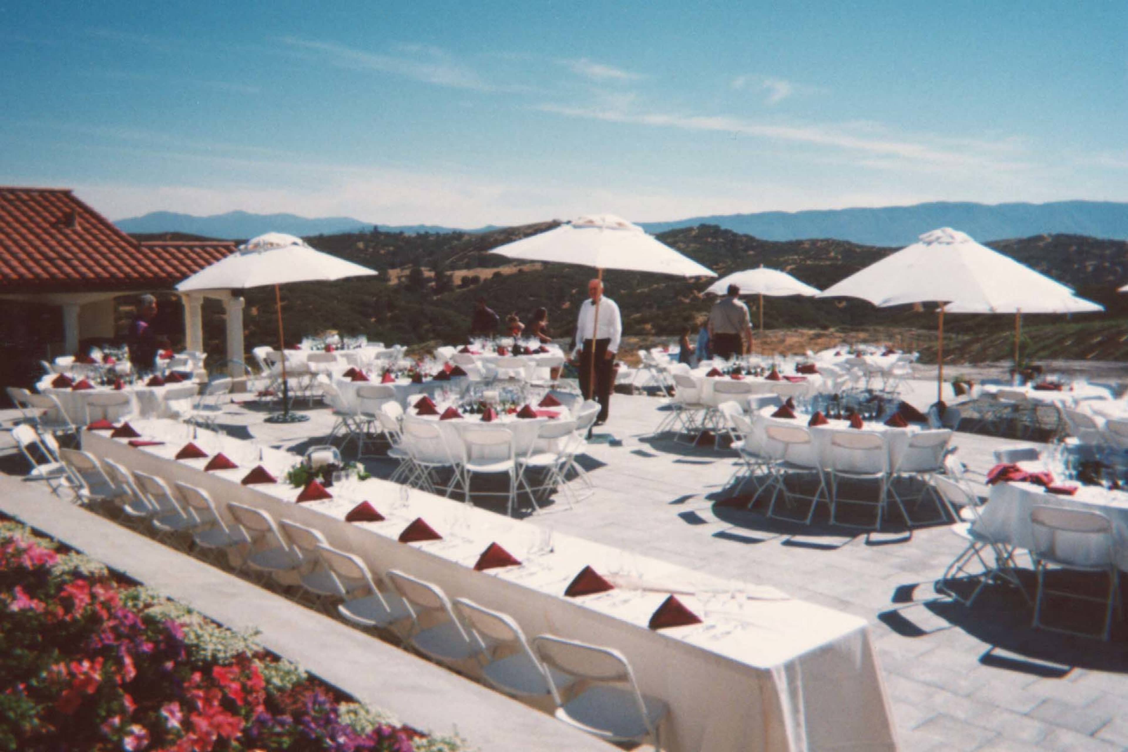 A large outdoor venue features multiple tables set for a dining event, with white chairs and umbrellas, overlooking a mountainous landscape.