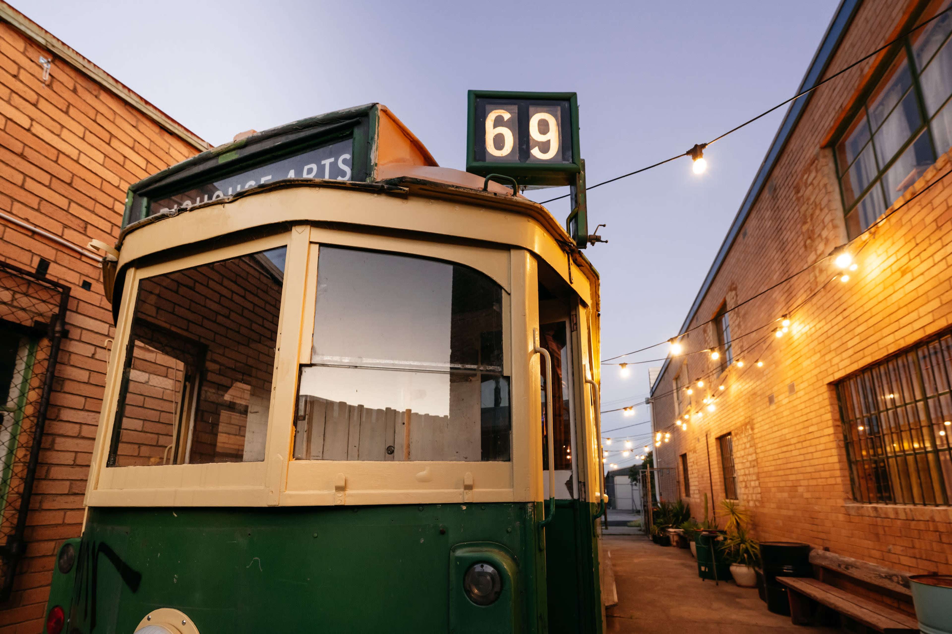 A vintage green tram with the number 69 displayed is parked in a narrow alley illuminated by string lights.