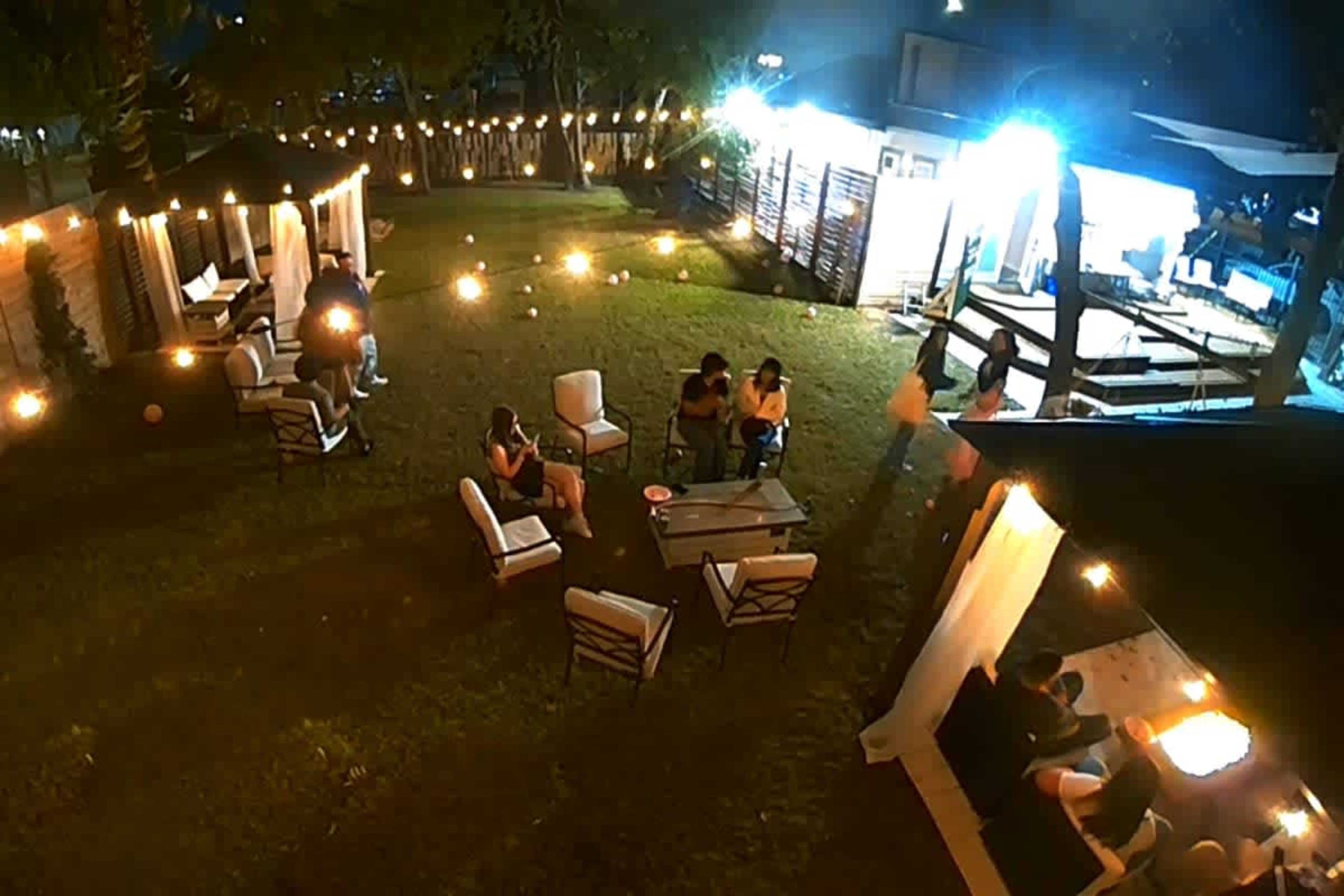 The image shows a gathering in a well-lit outdoor space with several people seated around tables and string lights illuminating the area.