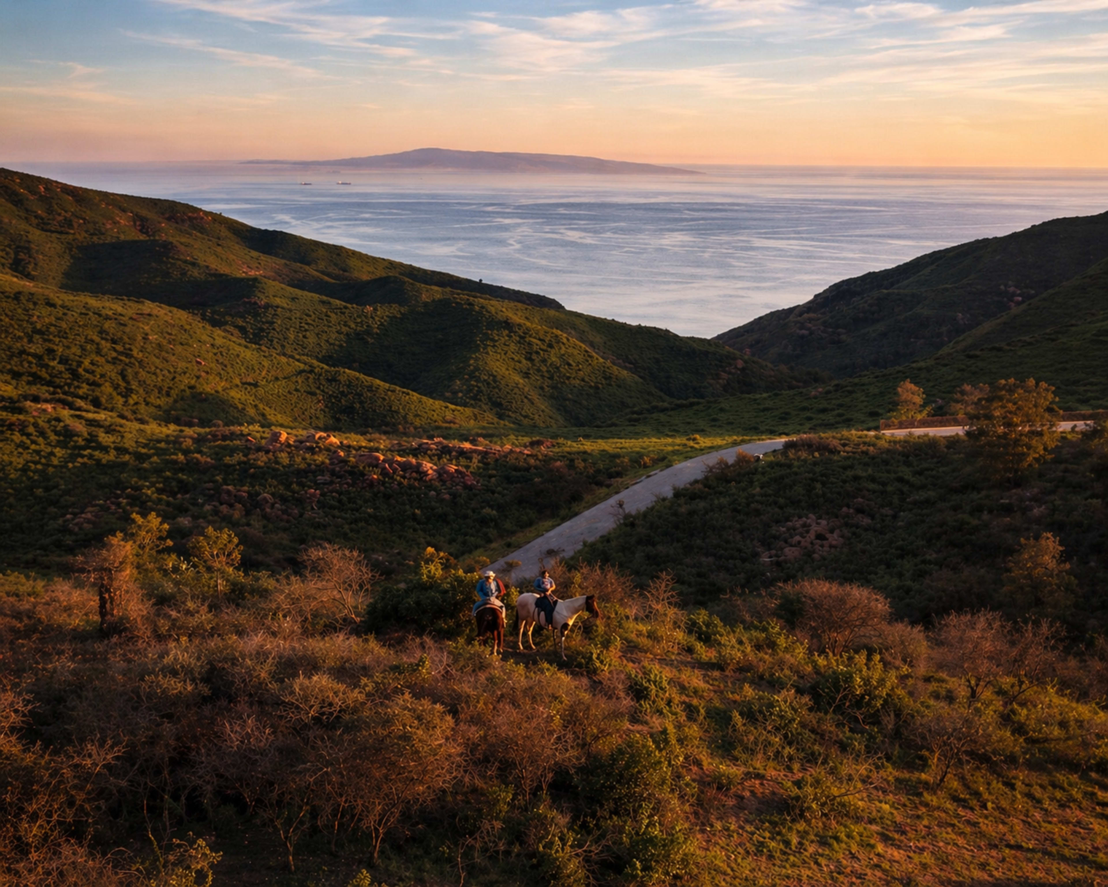 Two people on horseback stand on a hillside overlooking a body of water with distant islands at sunset.