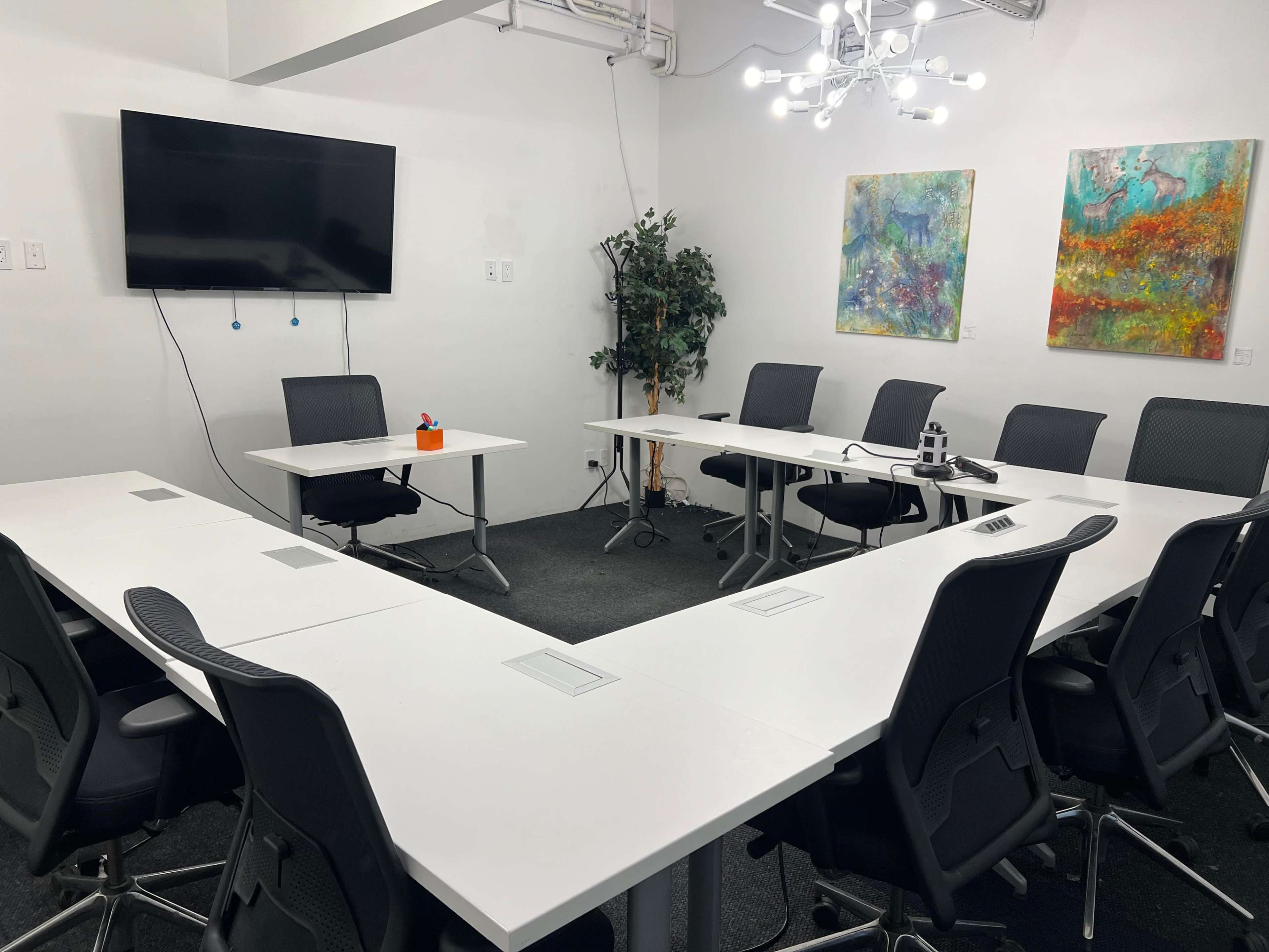 A conference room features a U-shaped table arrangement, a large wall-mounted screen, and two colorful abstract paintings.