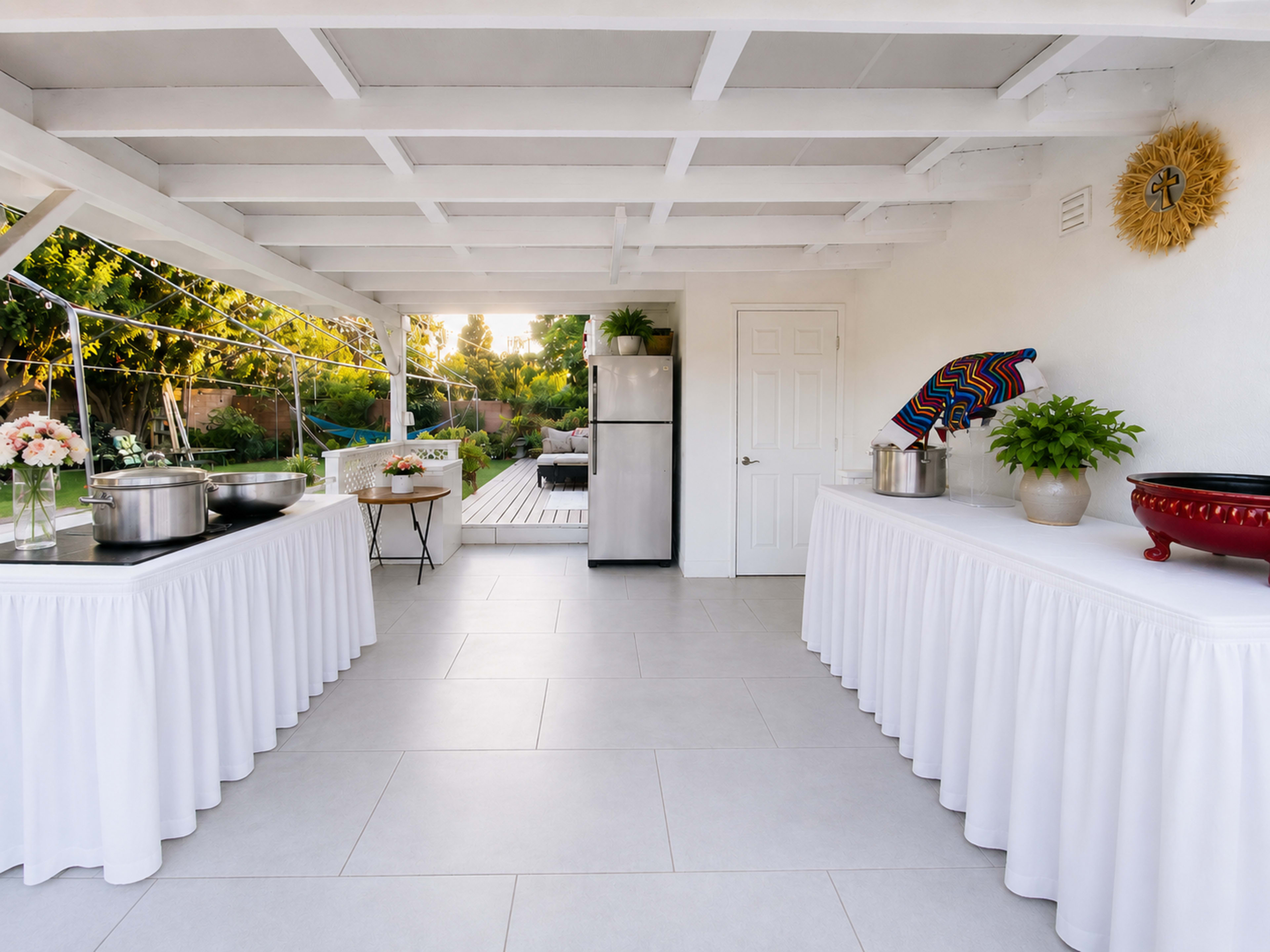 The image shows a covered outdoor space set up for an event, featuring two tables with white tablecloths, a large pot on one table, and a refrigerator in the background.
