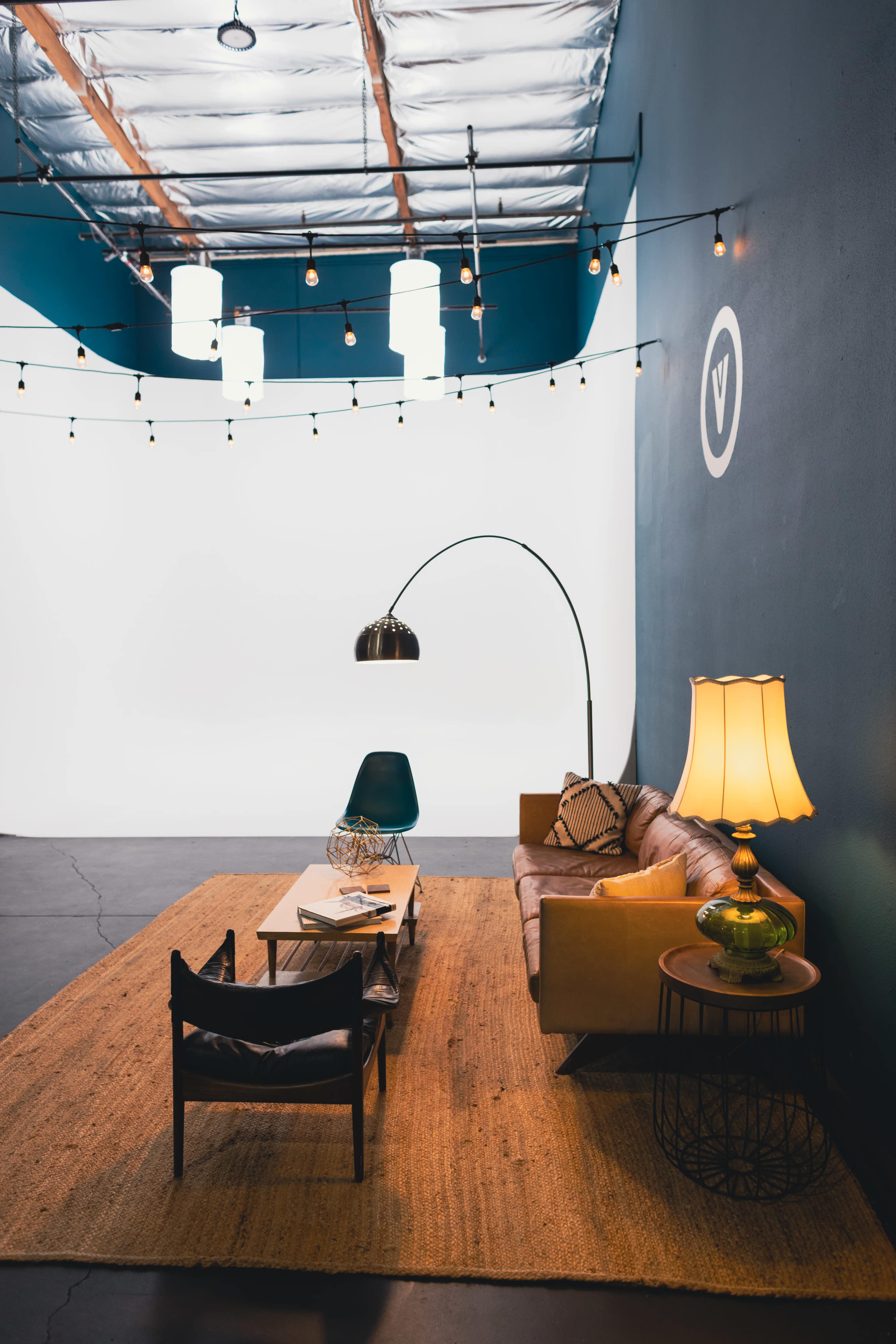 The image shows a modern interior living space featuring a brown leather sofa, a black lounge chair, a coffee table with magazines, a decorative lamp, and overhead string lights against a dark blue wall.