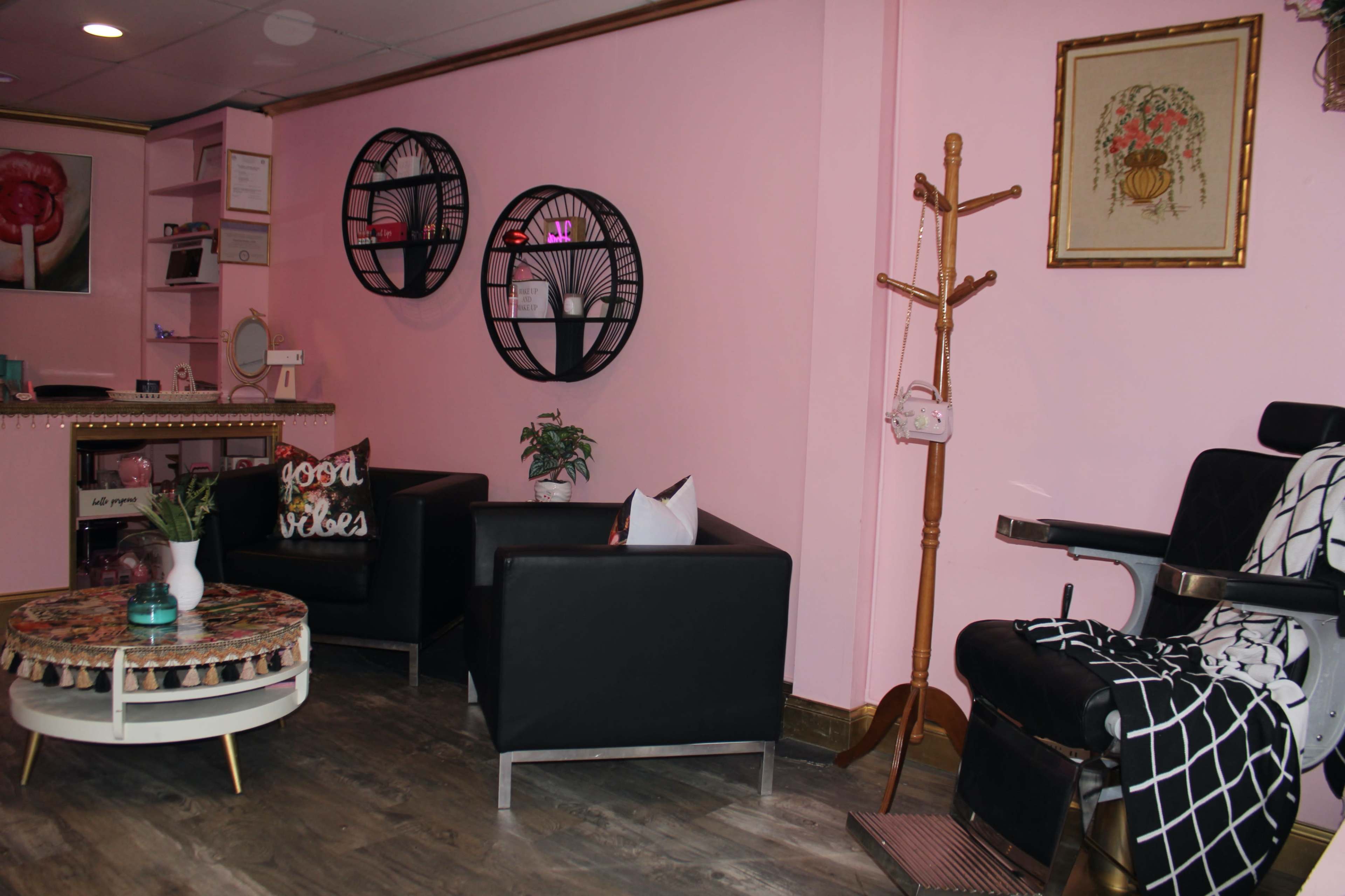 A cozy waiting area features black leather chairs, a round coffee table, and pink walls adorned with decorative wall art and plants.