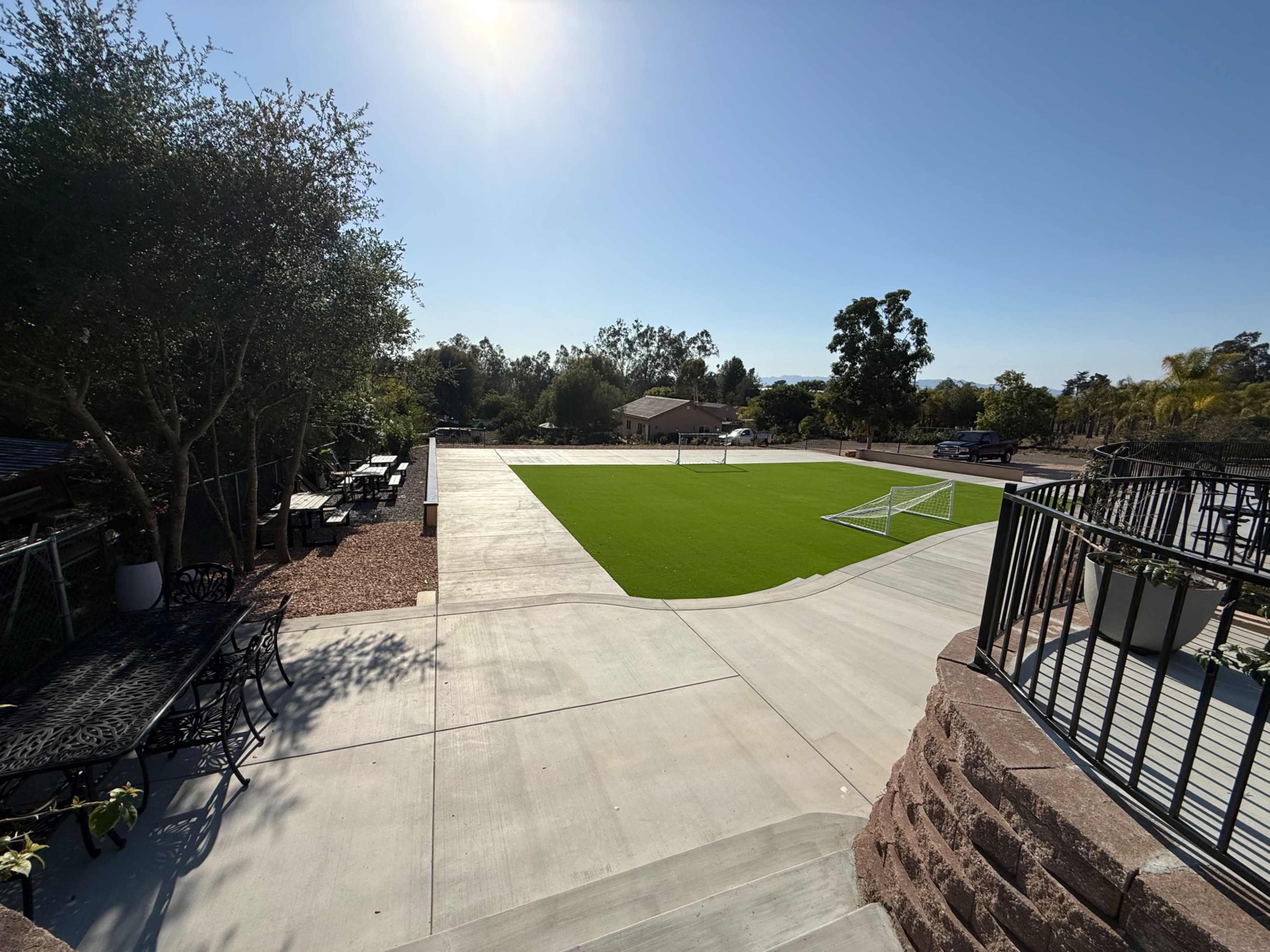 A bright, sunny view shows a large green soccer field with a goal, surrounded by trees and outdoor seating areas, leading down from a patio.
