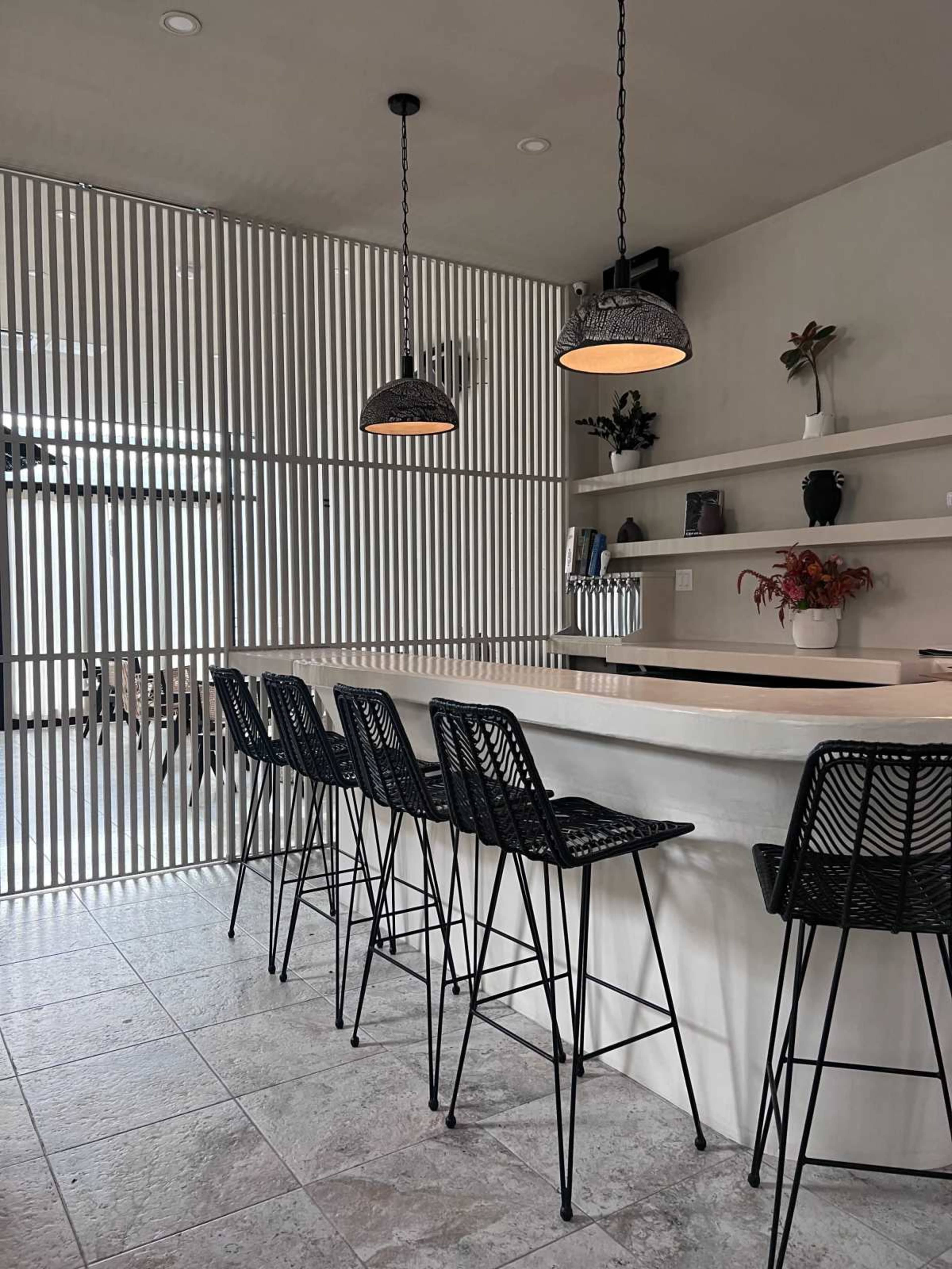 A modern kitchen features a minimalistic bar area with six black wire barstools, pendant lights, and a backdrop of slatted wood panels.