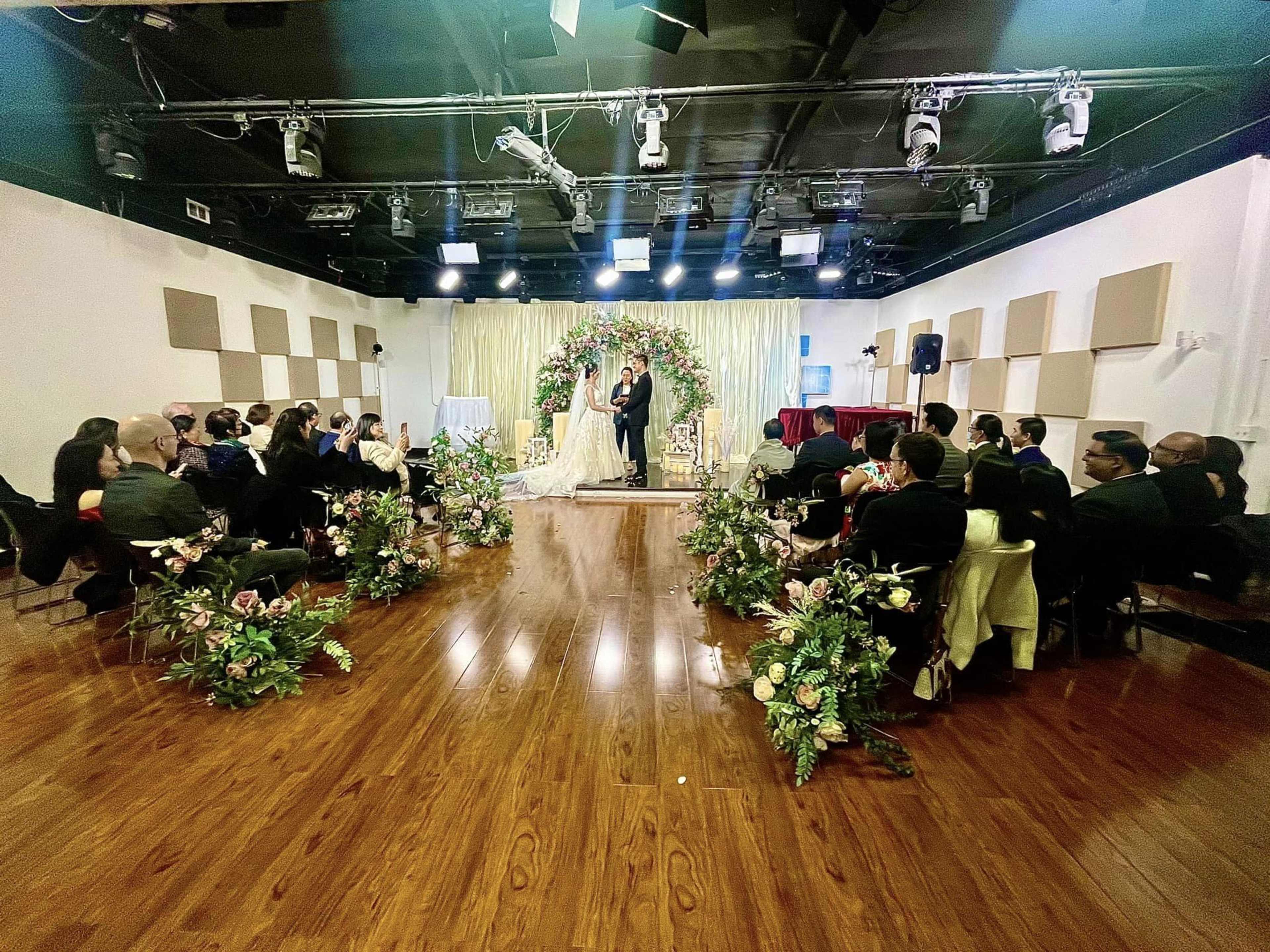 A couple stands in front of a floral arch while guests are seated in rows facing them in a decorated event space.