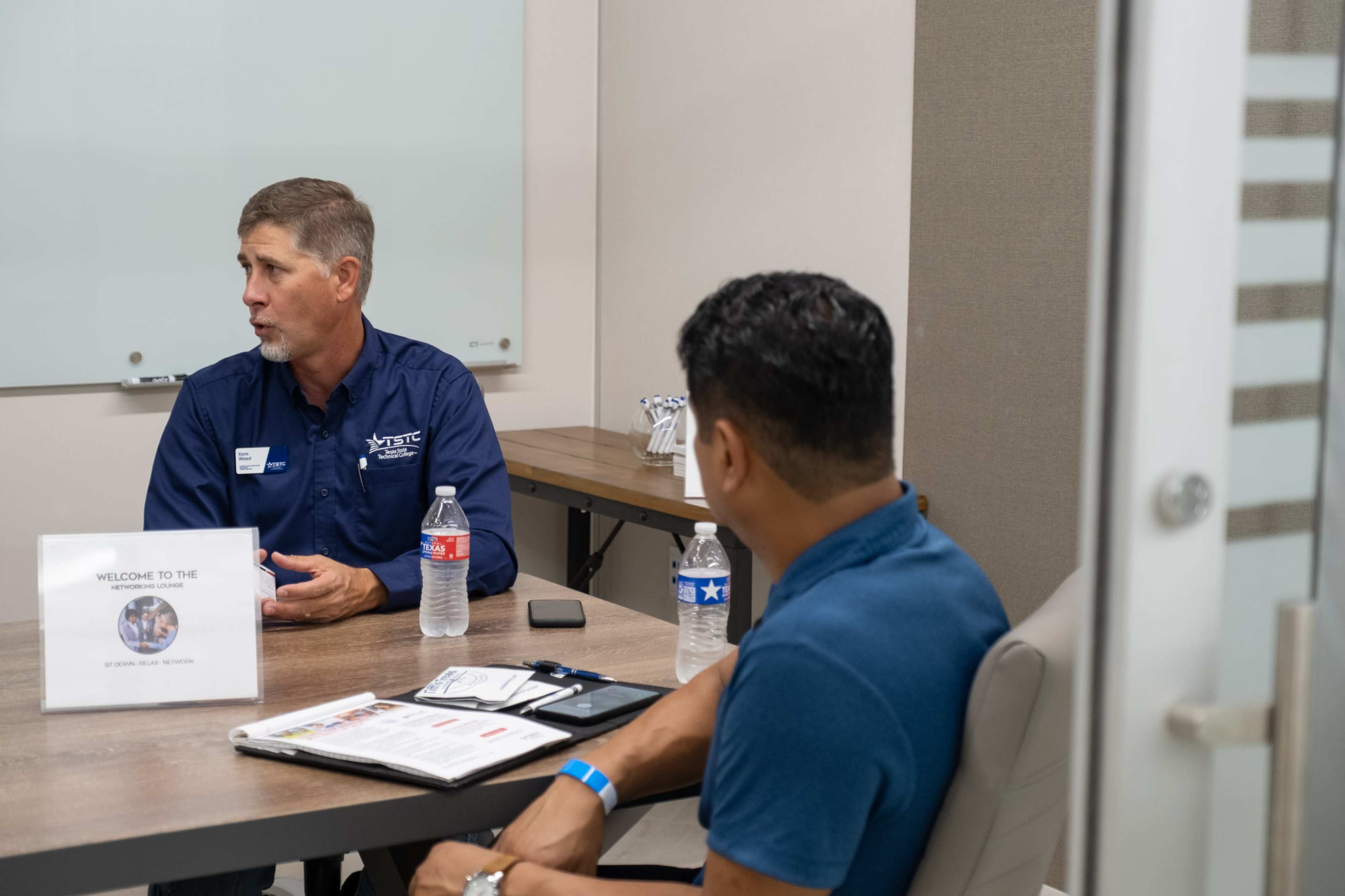 Two men are seated at a table in a meeting room, discussing while water bottles and a welcome sign are visible on the table.