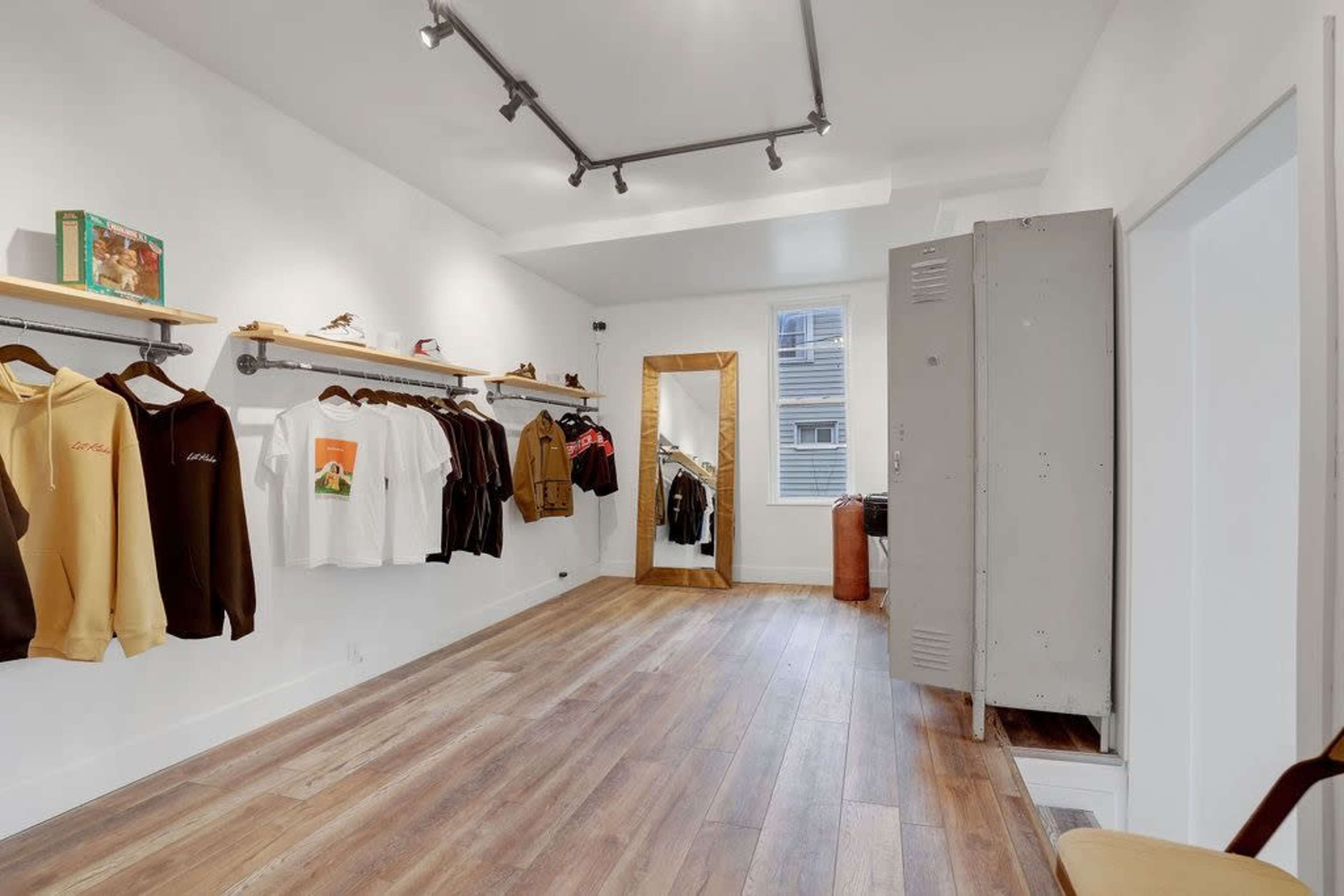 The image shows a minimalist clothing store with hanging garments on display, a large mirror, and a metal locker against the wall.