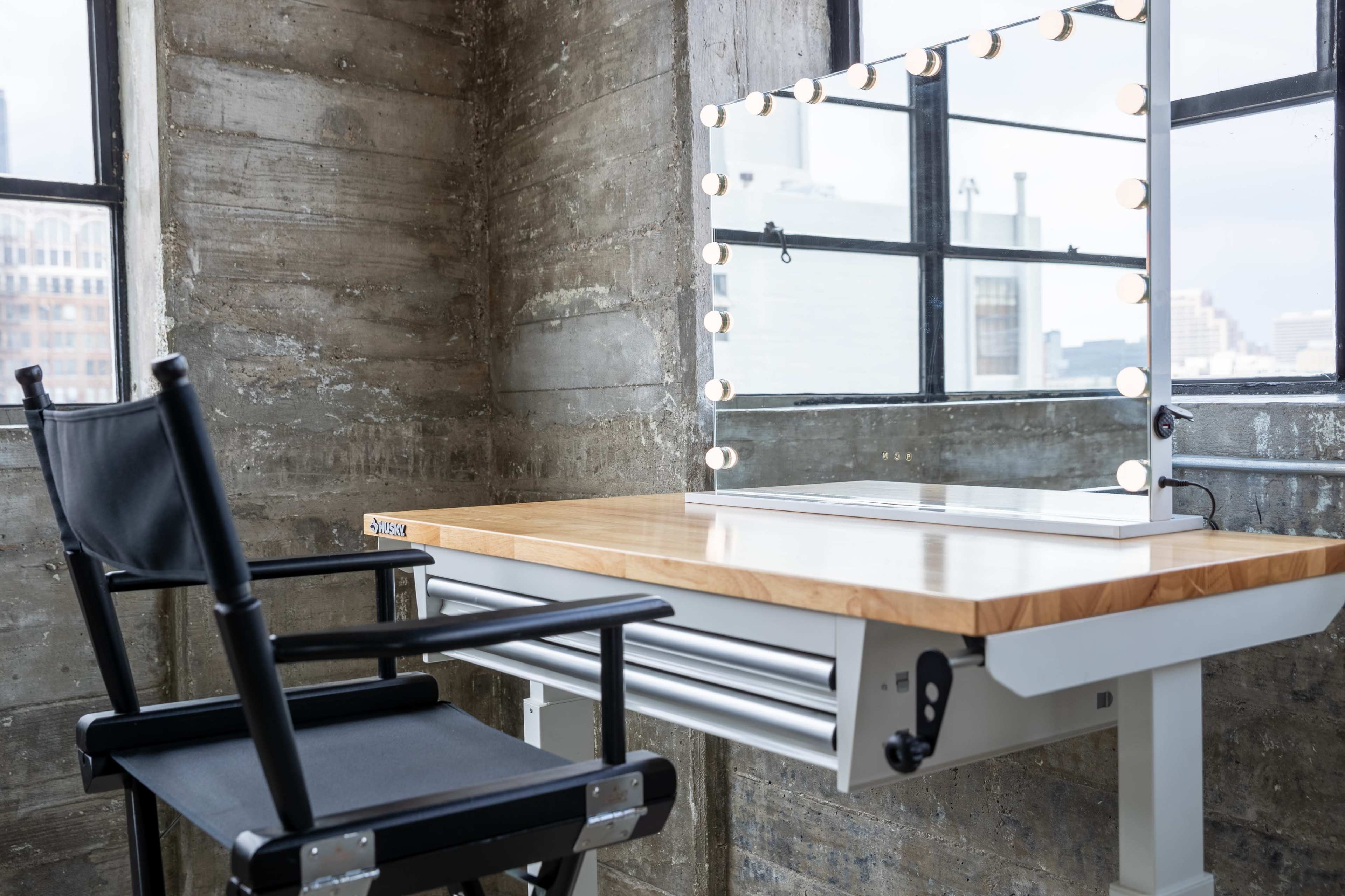 A black director's chair is positioned next to a wooden vanity table with a lighted mirror in a room featuring concrete walls and large windows.
