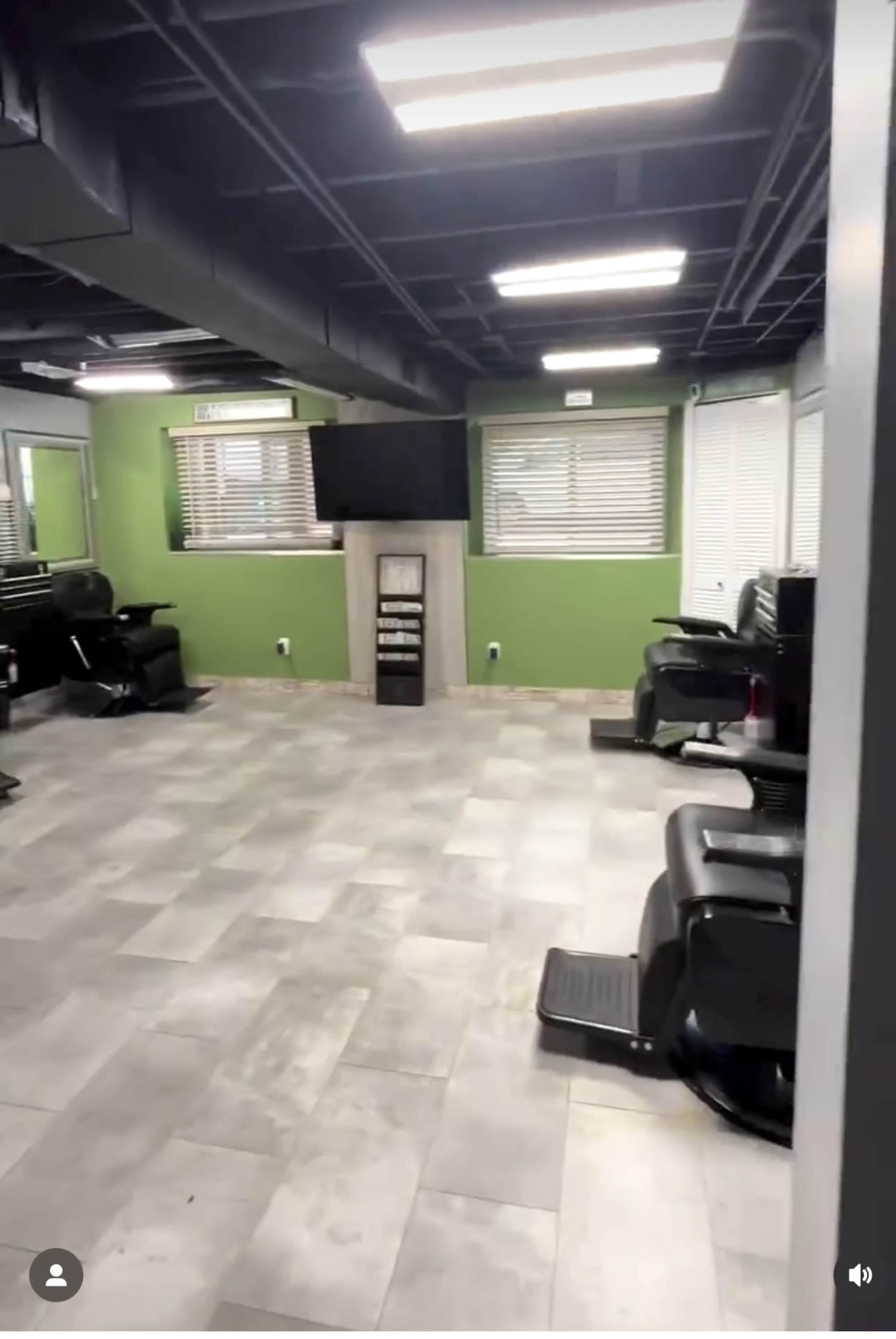A spacious barbershop with green walls, multiple black barber chairs, and a large television mounted on the wall.