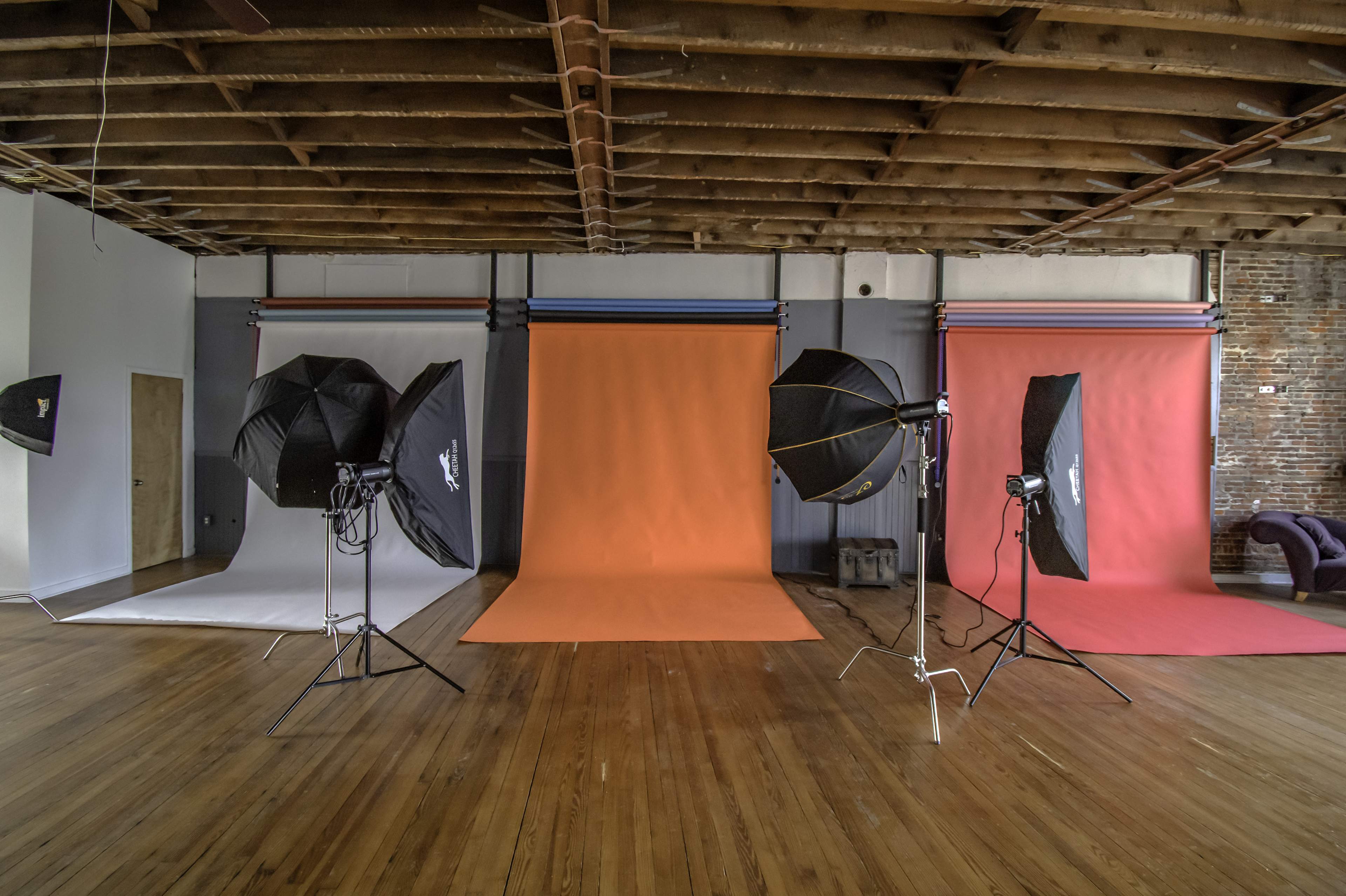A photography studio with three backdrop setups, including a gray, orange, and light blue background, along with multiple lighting equipment positioned around the space.