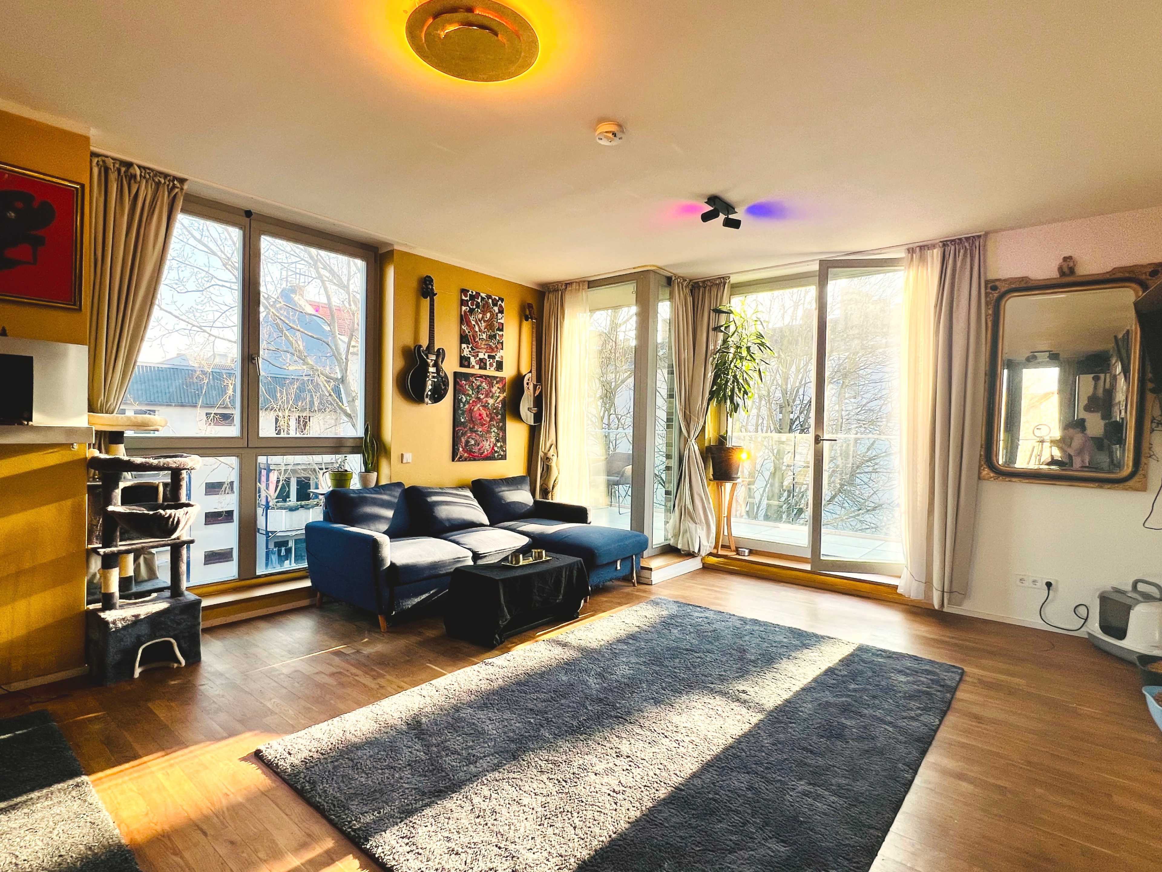The image shows a brightly lit living room with large windows, a blue couch, a coffee table, and decorative artwork on the walls.