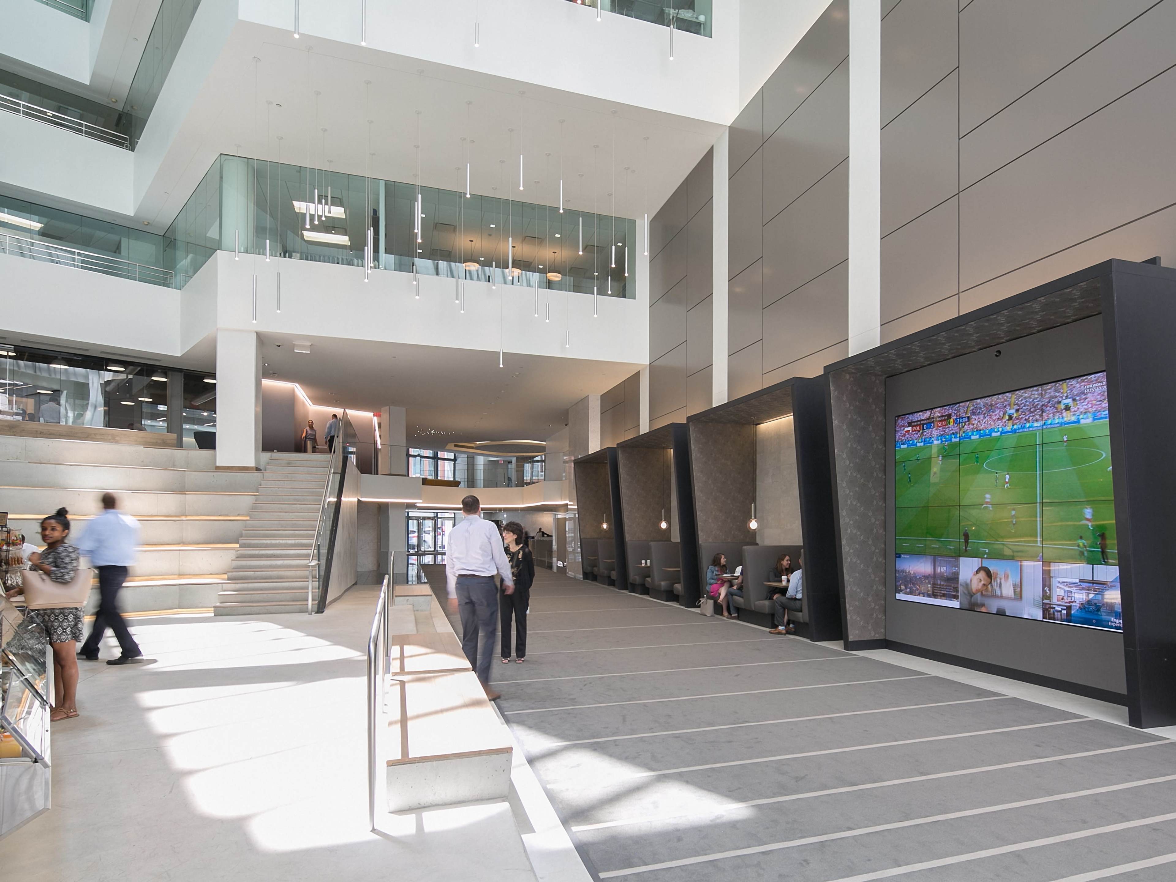 The image shows a modern, open atrium with a staircase leading to an upper level, featuring large screens displaying a sports event along one wall.
