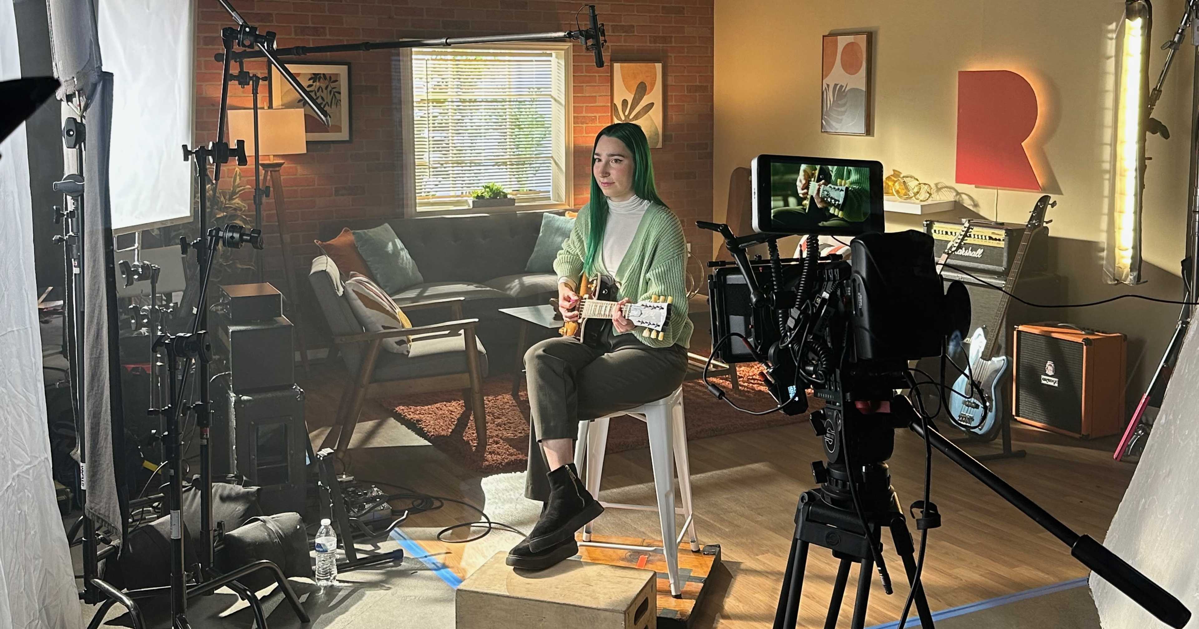 A musician sits on a stool in a well-lit studio, playing an electric guitar while being filmed by a camera on a tripod.