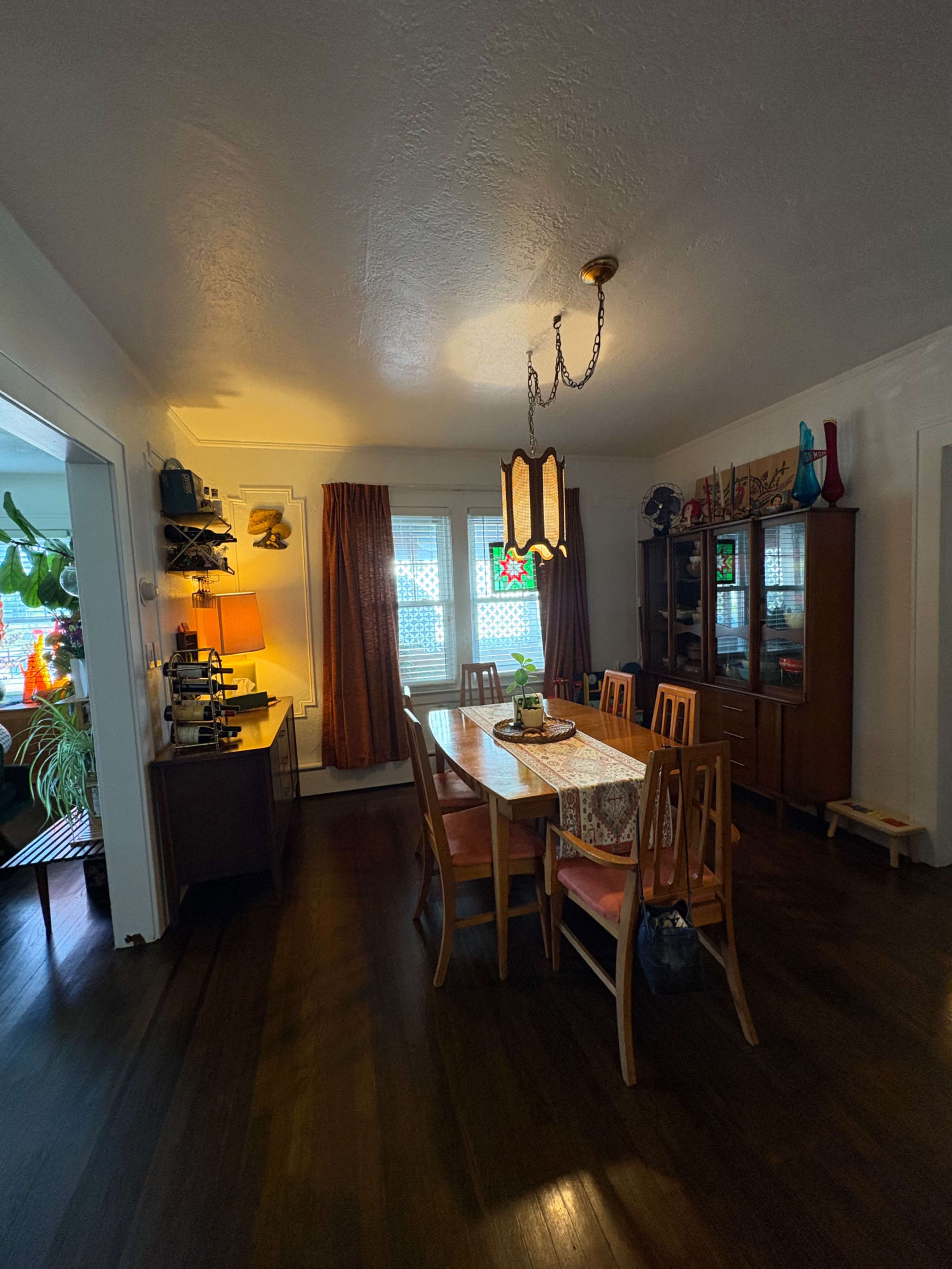 The image shows a dining room with a wooden table surrounded by chairs, a cabinet displaying various items, and warm lighting from a ceiling fixture.