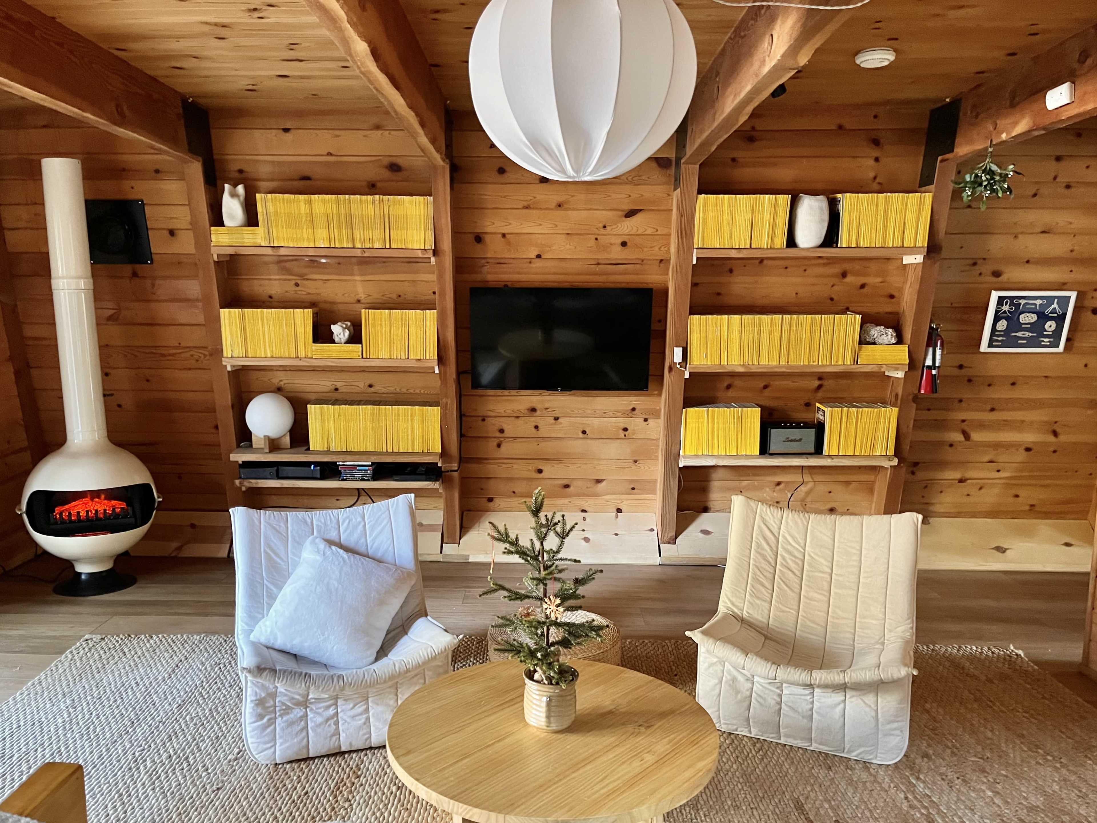 The image depicts a cozy wooden interior with two light-colored chairs, a small potted tree, a round wooden table, and yellow bookcases filled with books.