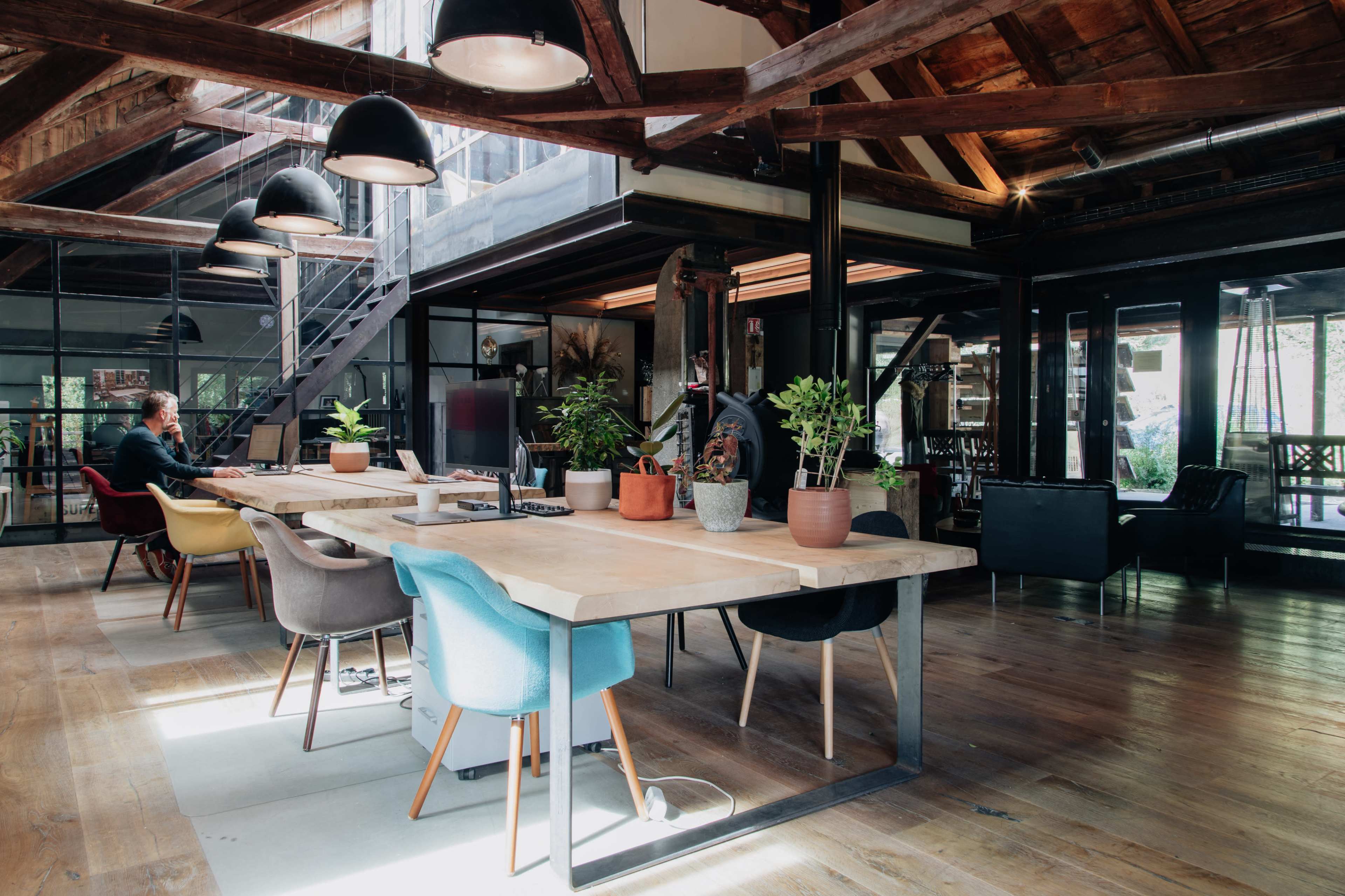 The image depicts a spacious office interior with wooden beams, multiple seating areas, and large windows allowing natural light to stream in.