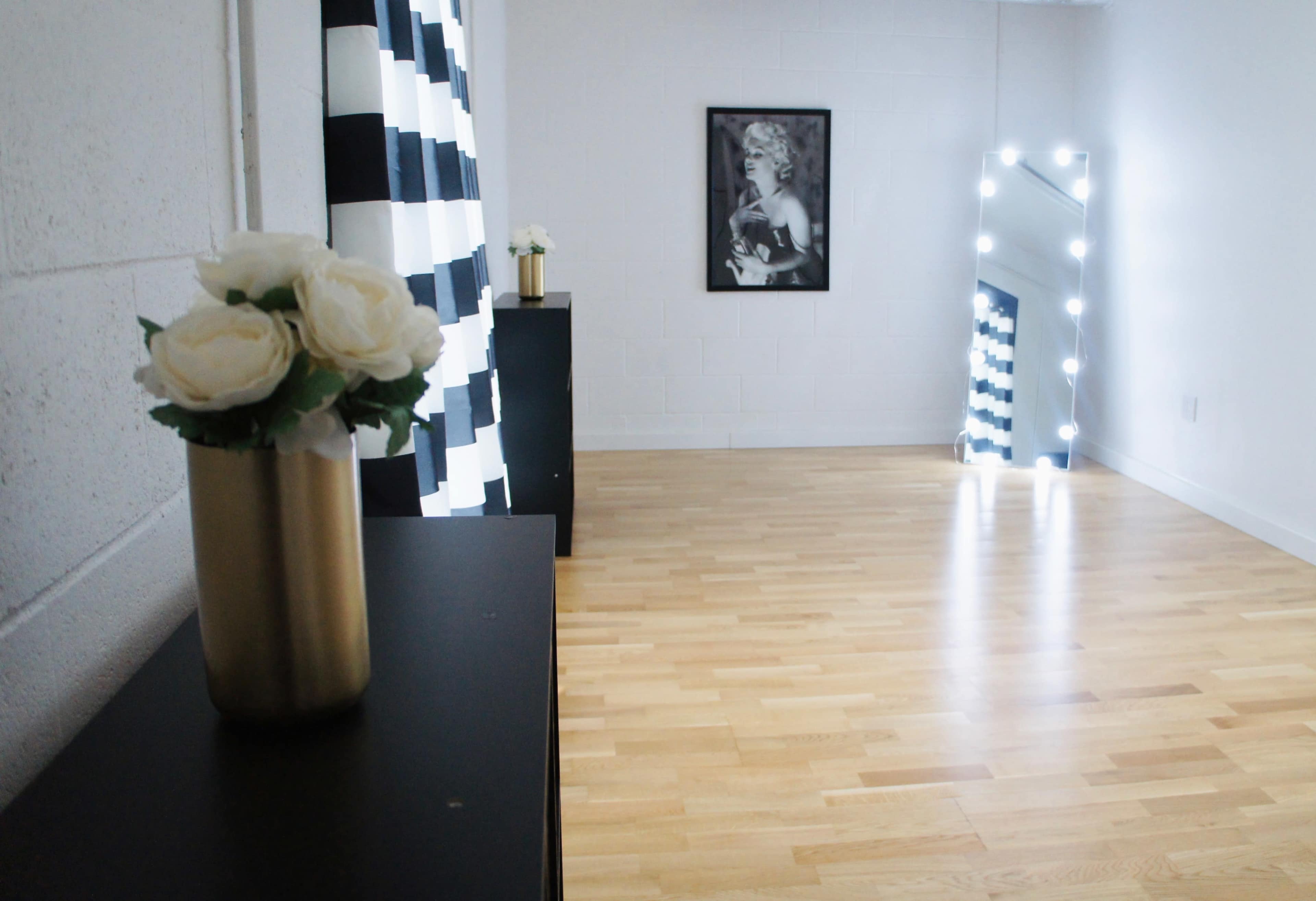 A bright room with wooden flooring, a black dresser, a mirror with lights, and a black-and-white striped curtain.