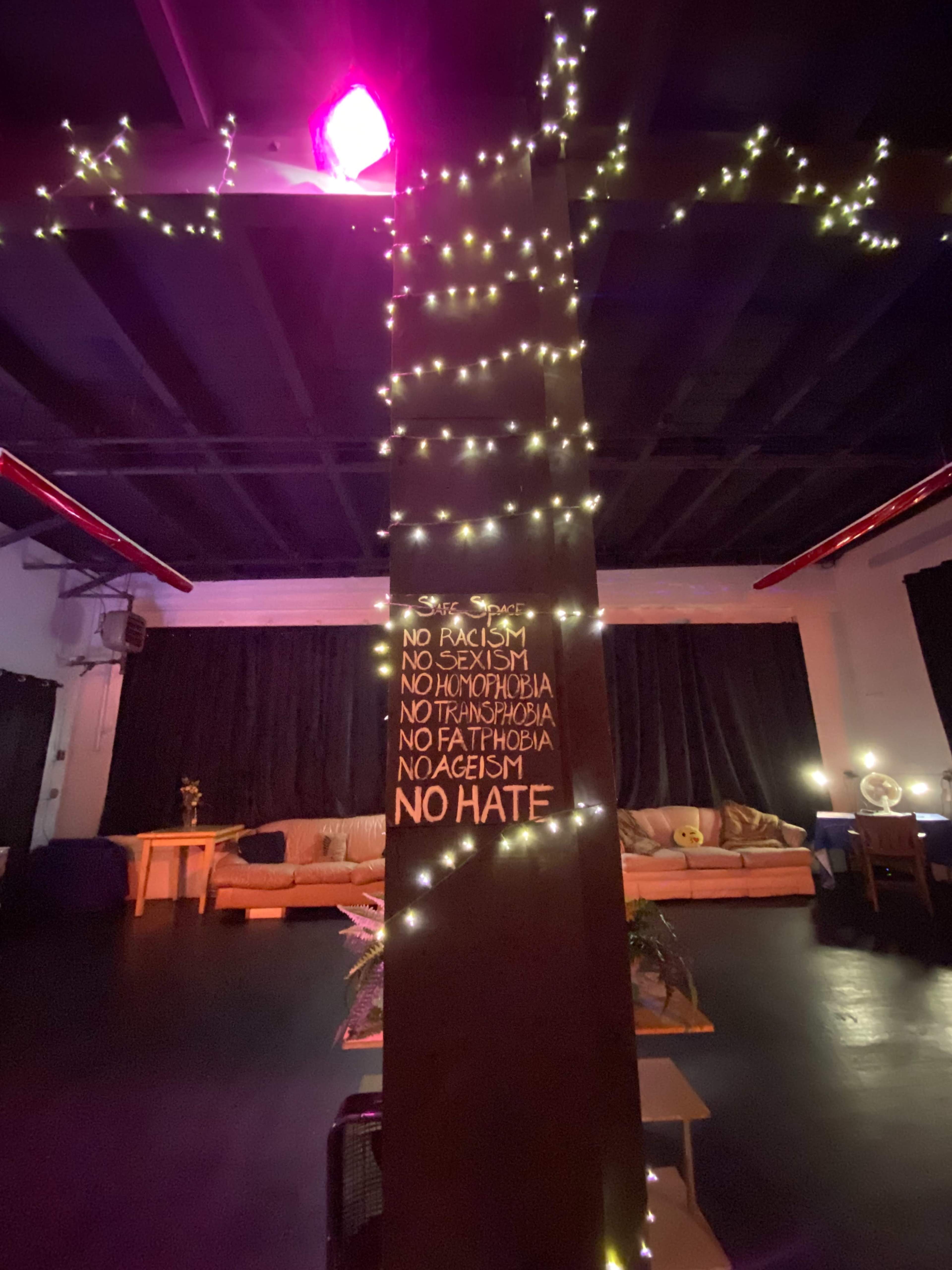 A dimly lit interior with a wooden pillar wrapped in string lights, featuring a sign that lists various forms of discrimination, including racism and sexism, alongside the phrase "NO HATE."