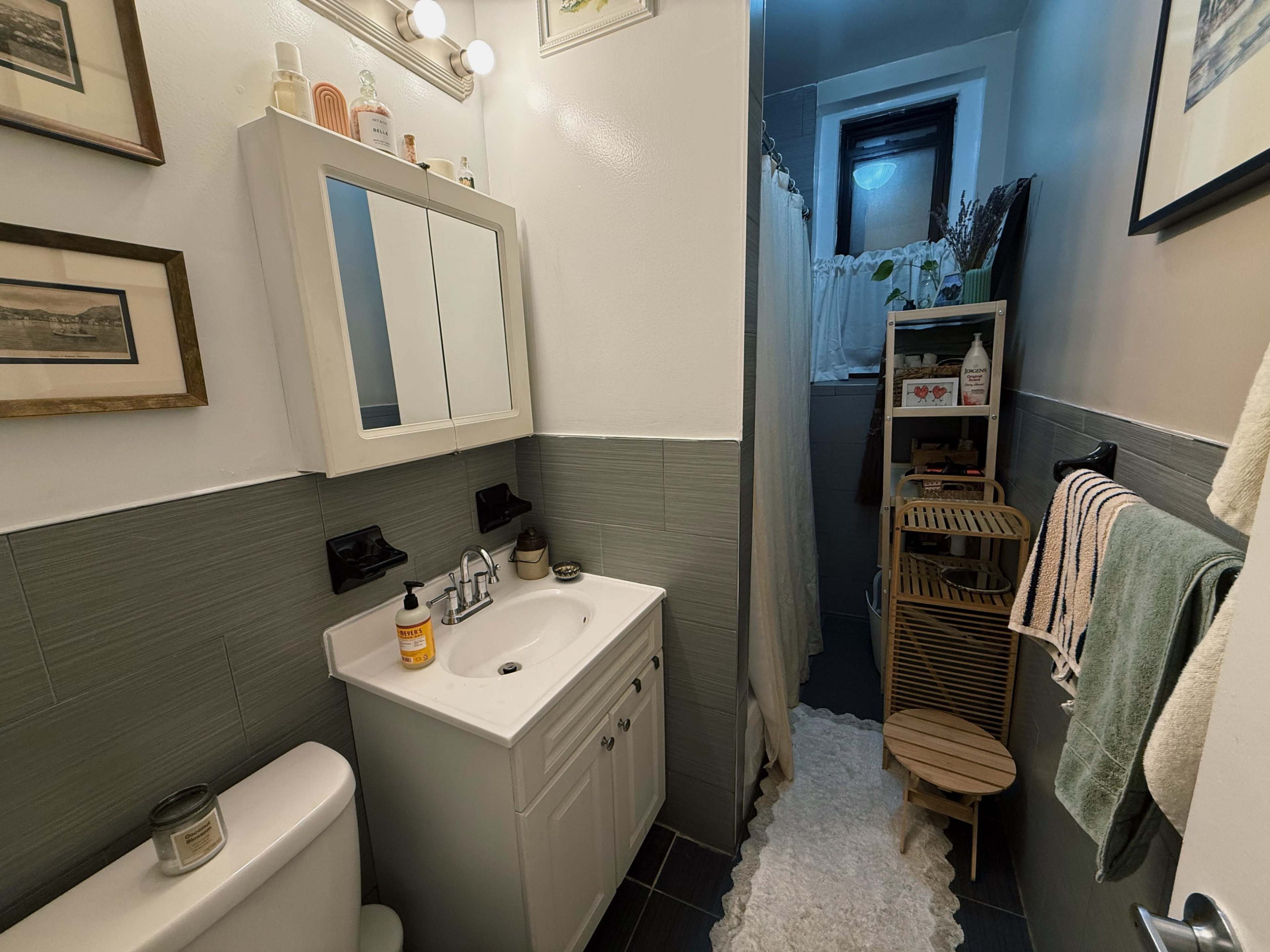 The image shows a narrow bathroom featuring a sink with a mirror above, a shower curtain in the corner, and a wooden shelf holding toiletries and towels.