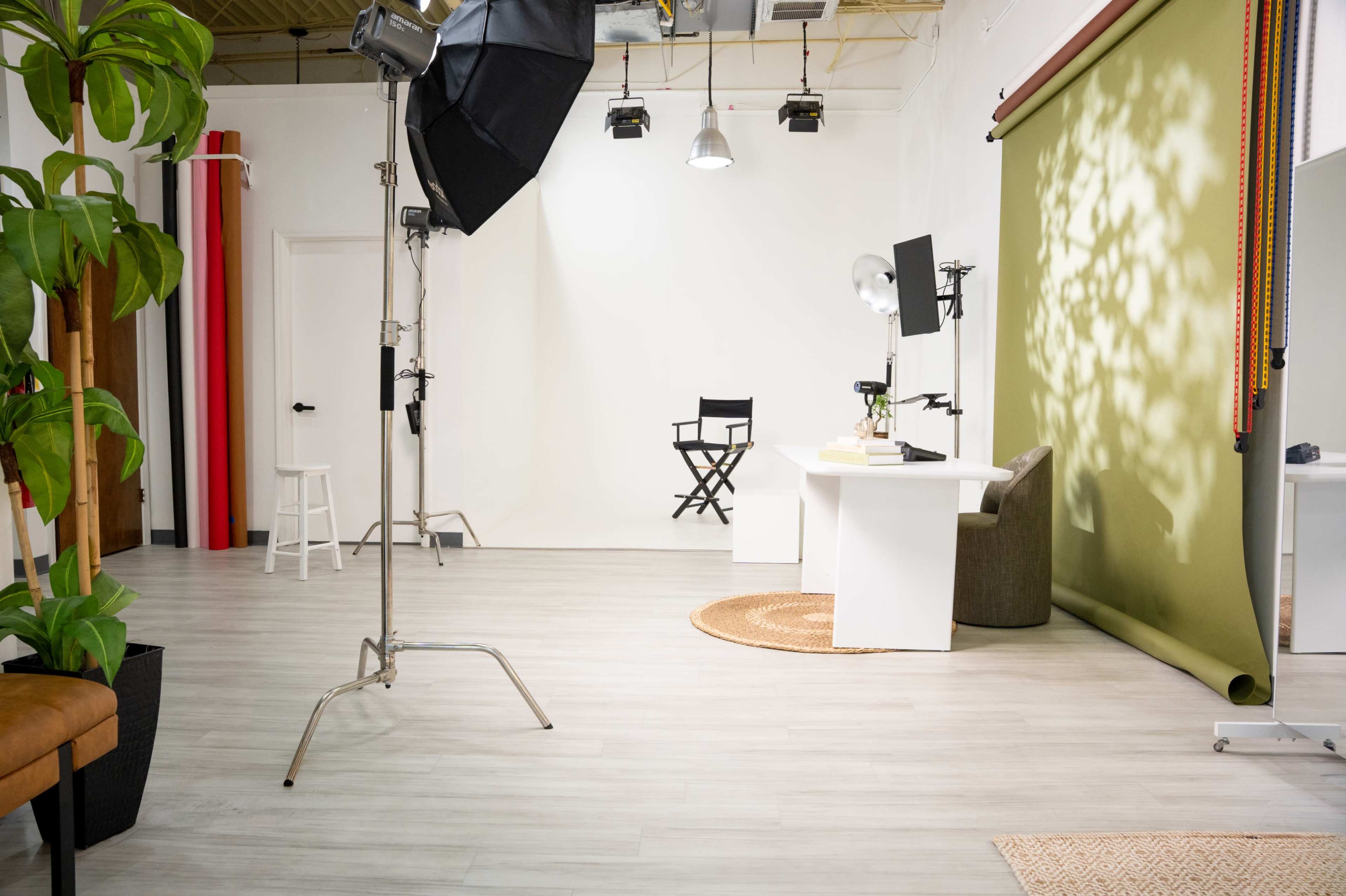 The image shows a photography studio with lighting equipment, a green backdrop, a white desk, a chair, and a potted plant.