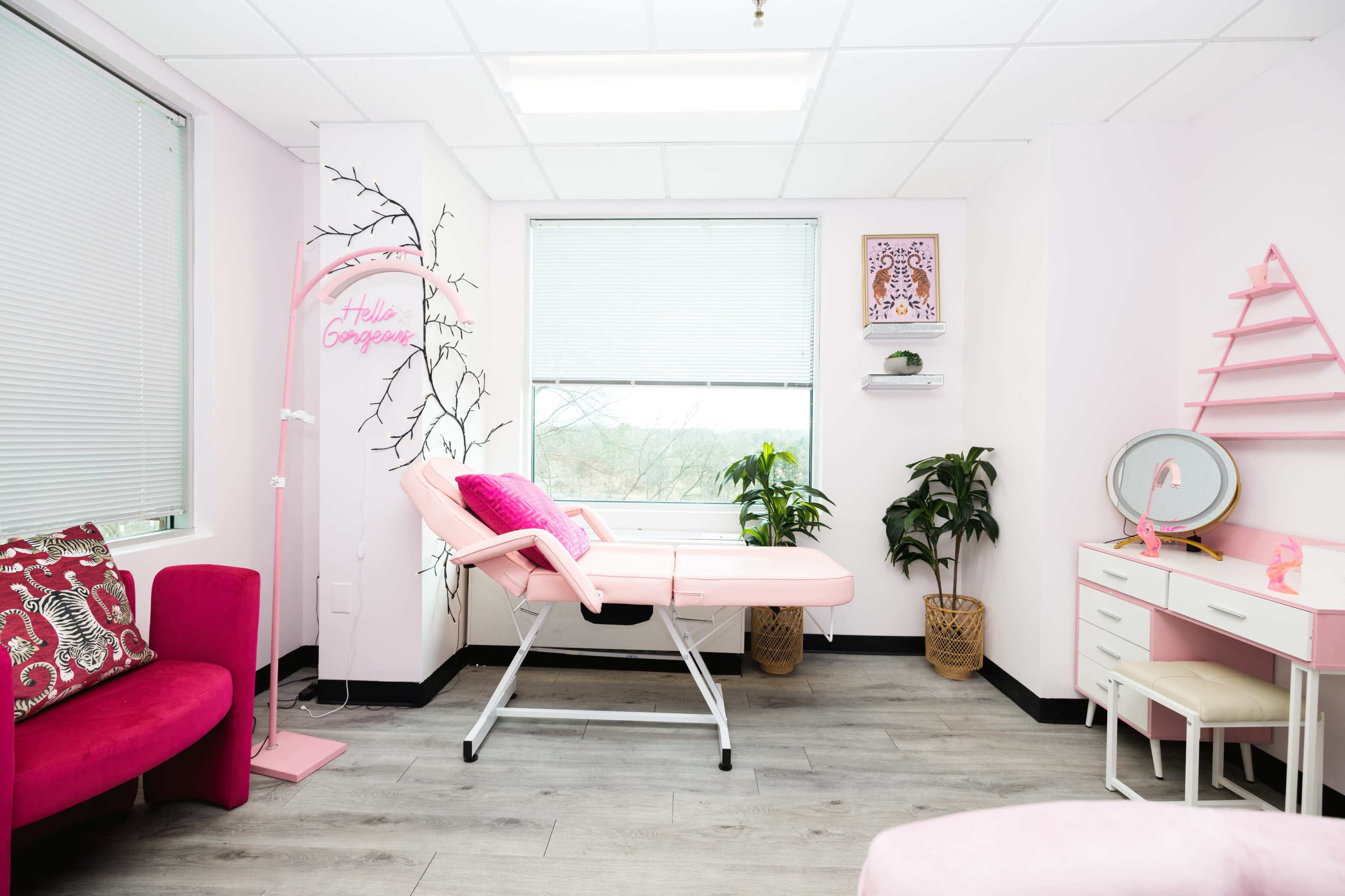 A light pink aesthetic room featuring a treatment bed, a small desk, and decorative plants.