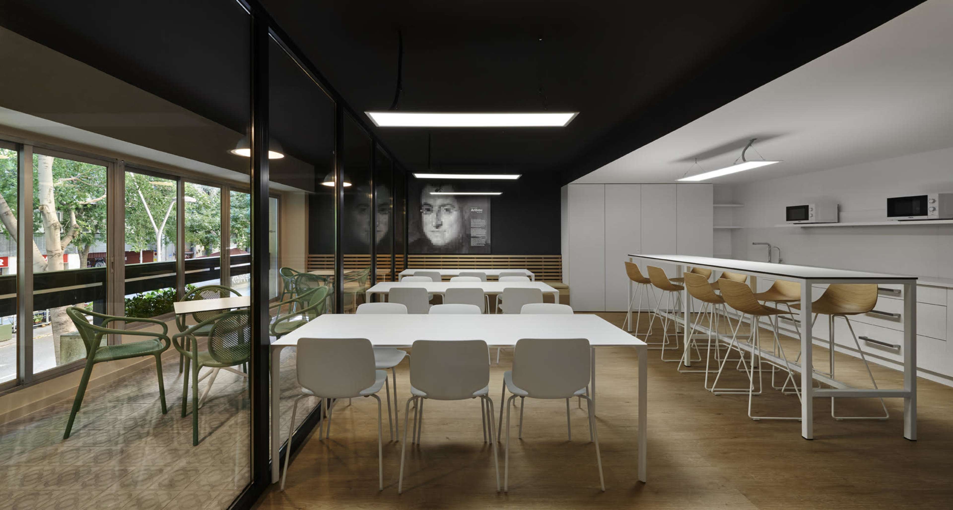 The image shows a modern cafeteria with white tables and chairs, a bar area with tall stools, and large windows overlooking a green outdoor space.