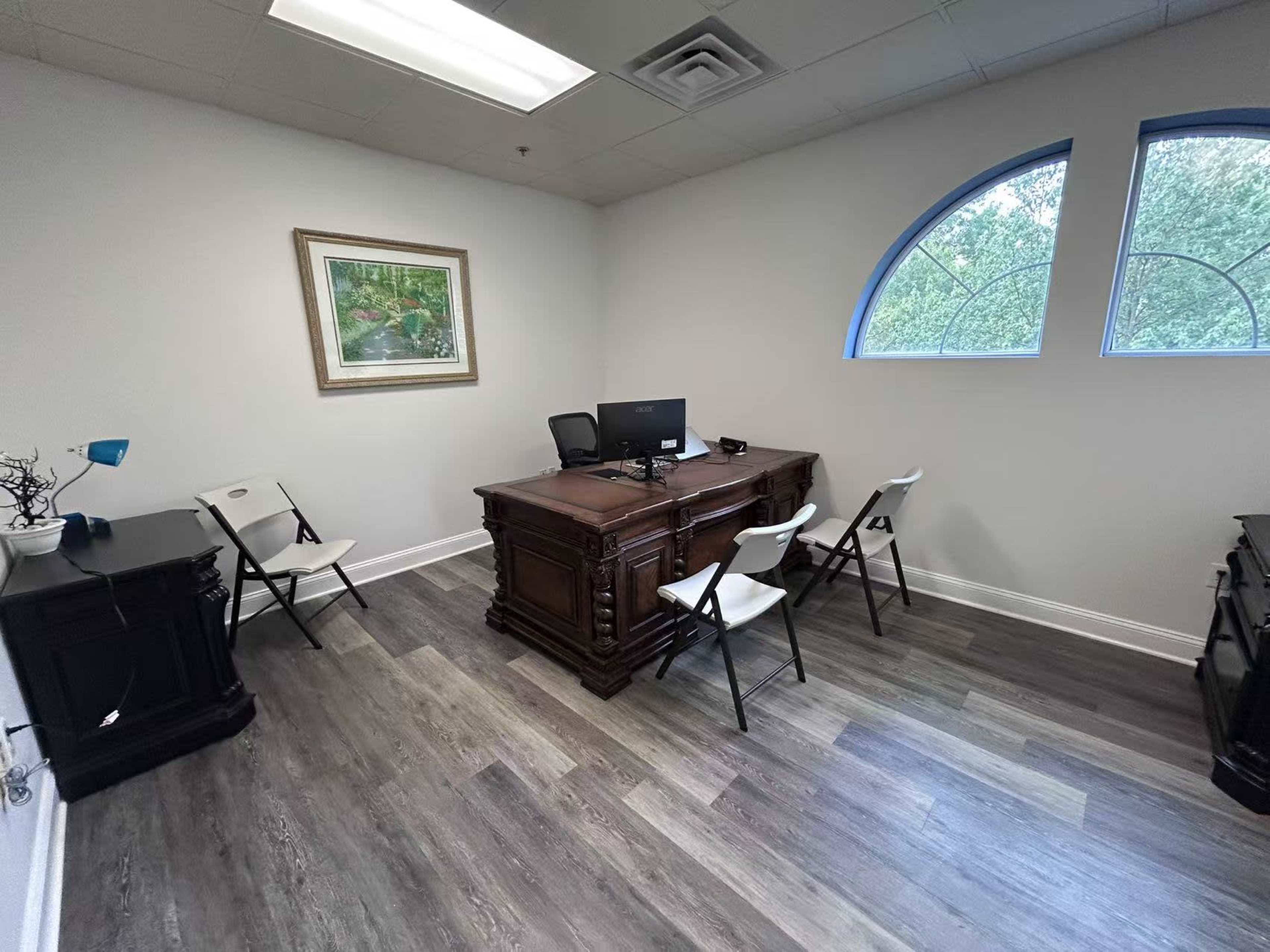 The image shows a sparsely furnished office with a wooden desk, a computer, and four white folding chairs arranged around the room.