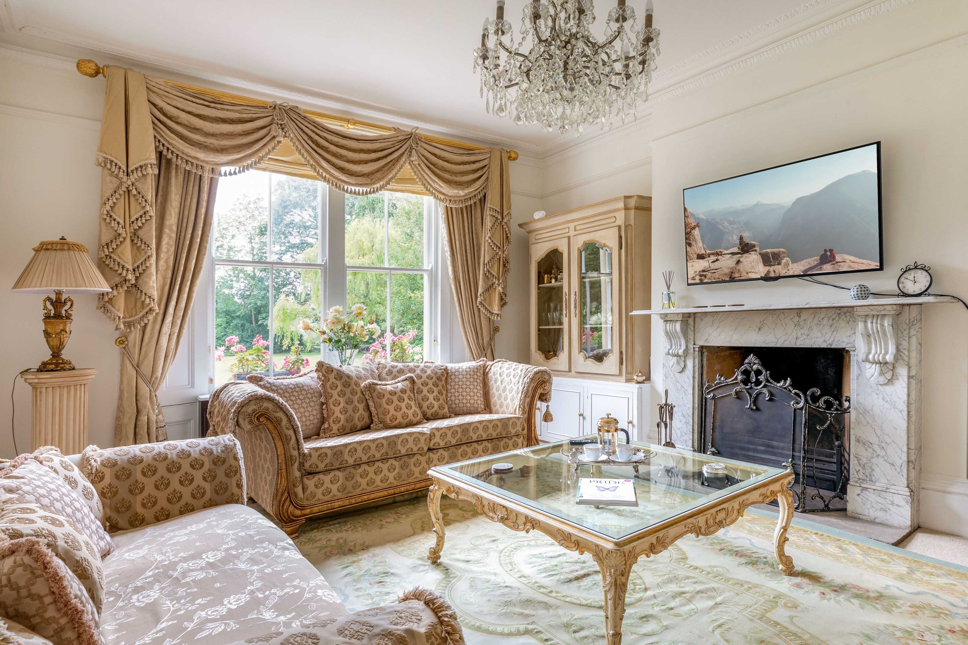 An elegantly furnished living room with a crystal chandelier, ornate furniture, and large windows overlooking a garden.