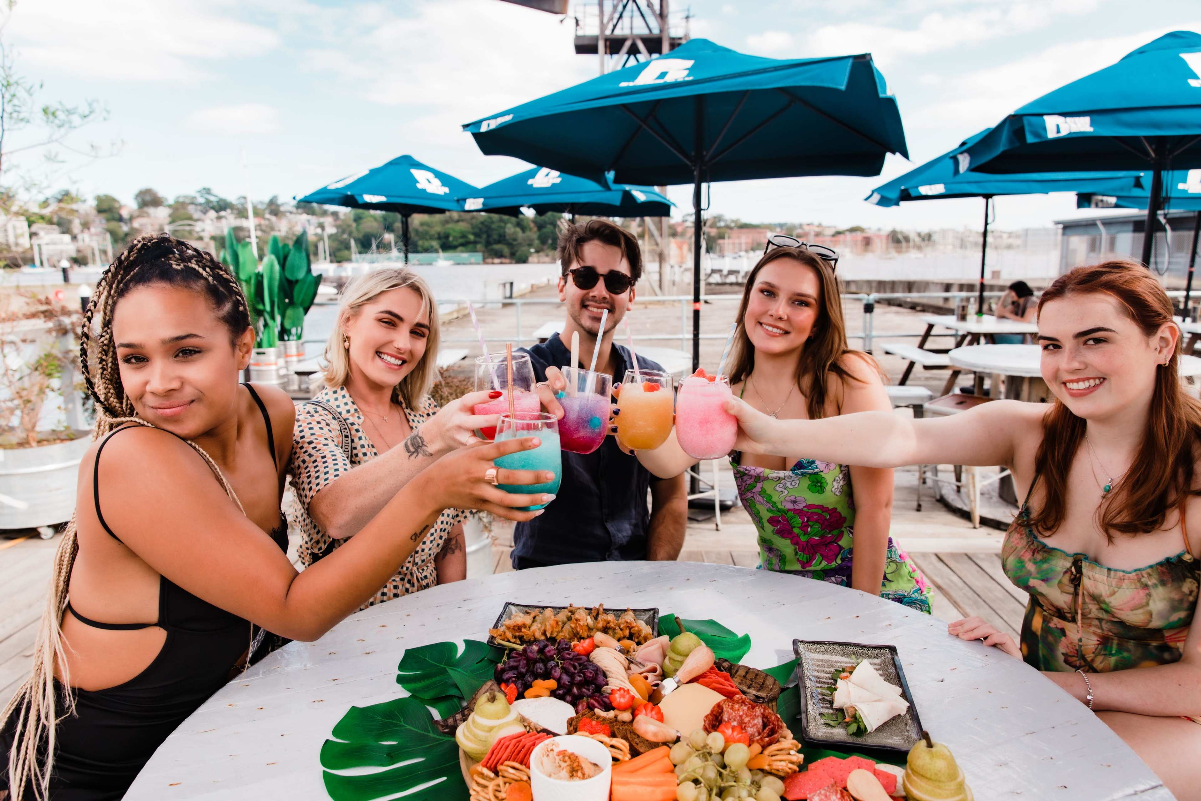 A group of five people is seated at a table outdoors, holding colorful drinks while surrounded by a spread of food and blue umbrellas.