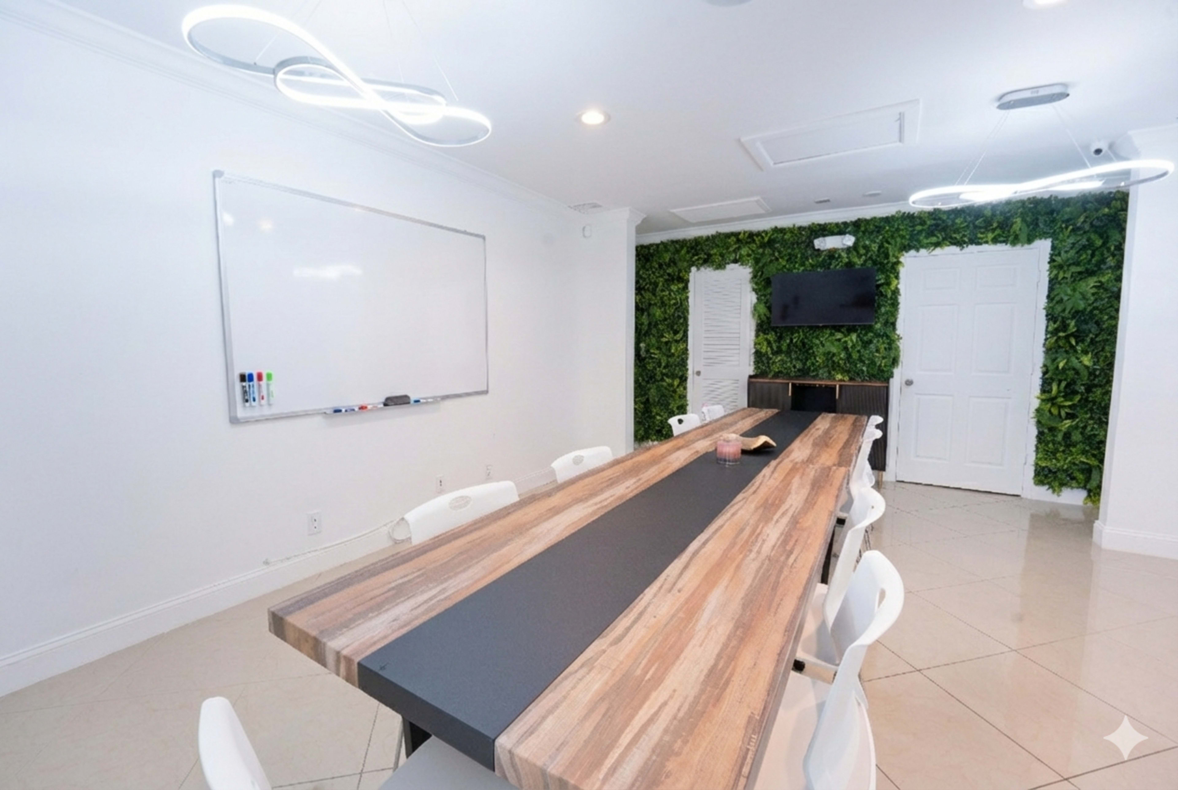 A conference room features a long wooden table, white chairs, a whiteboard on the wall, and a green plant wall with a TV mounted above.