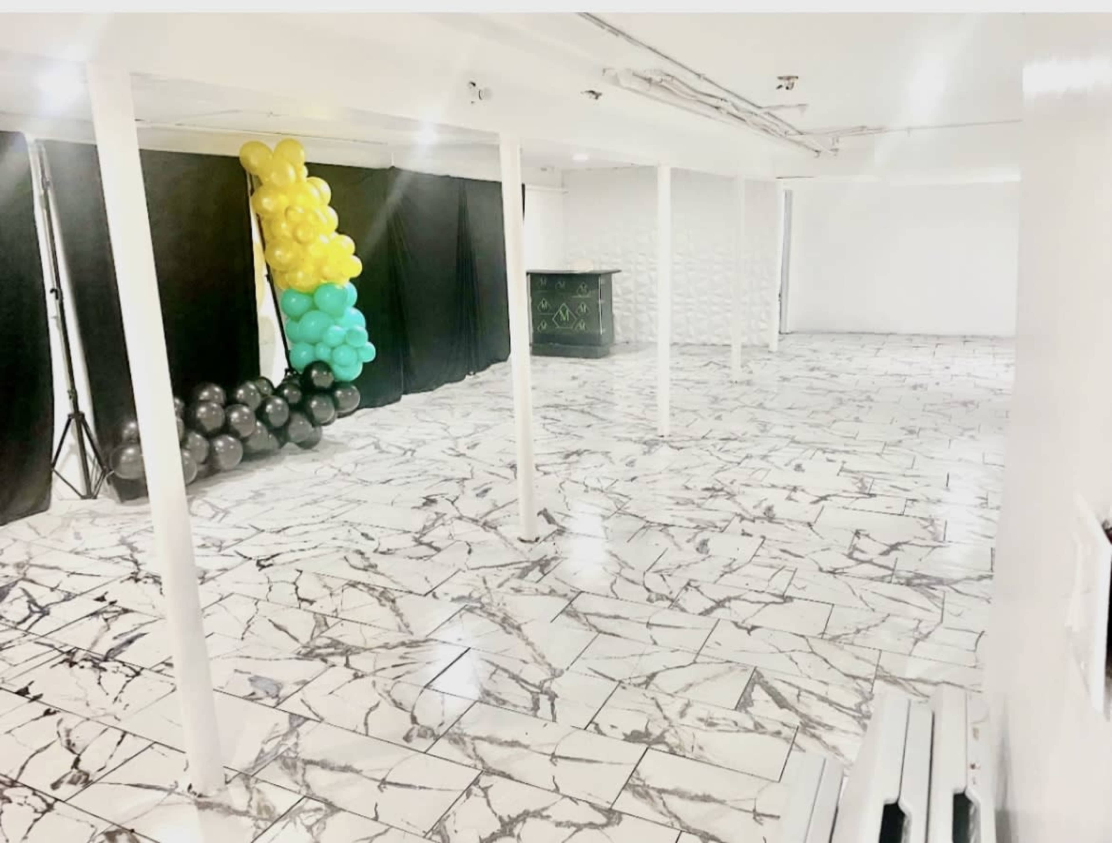 A spacious interior room with marble-patterned flooring, black curtains, a green bar, and balloons in yellow and green arranged in a corner.