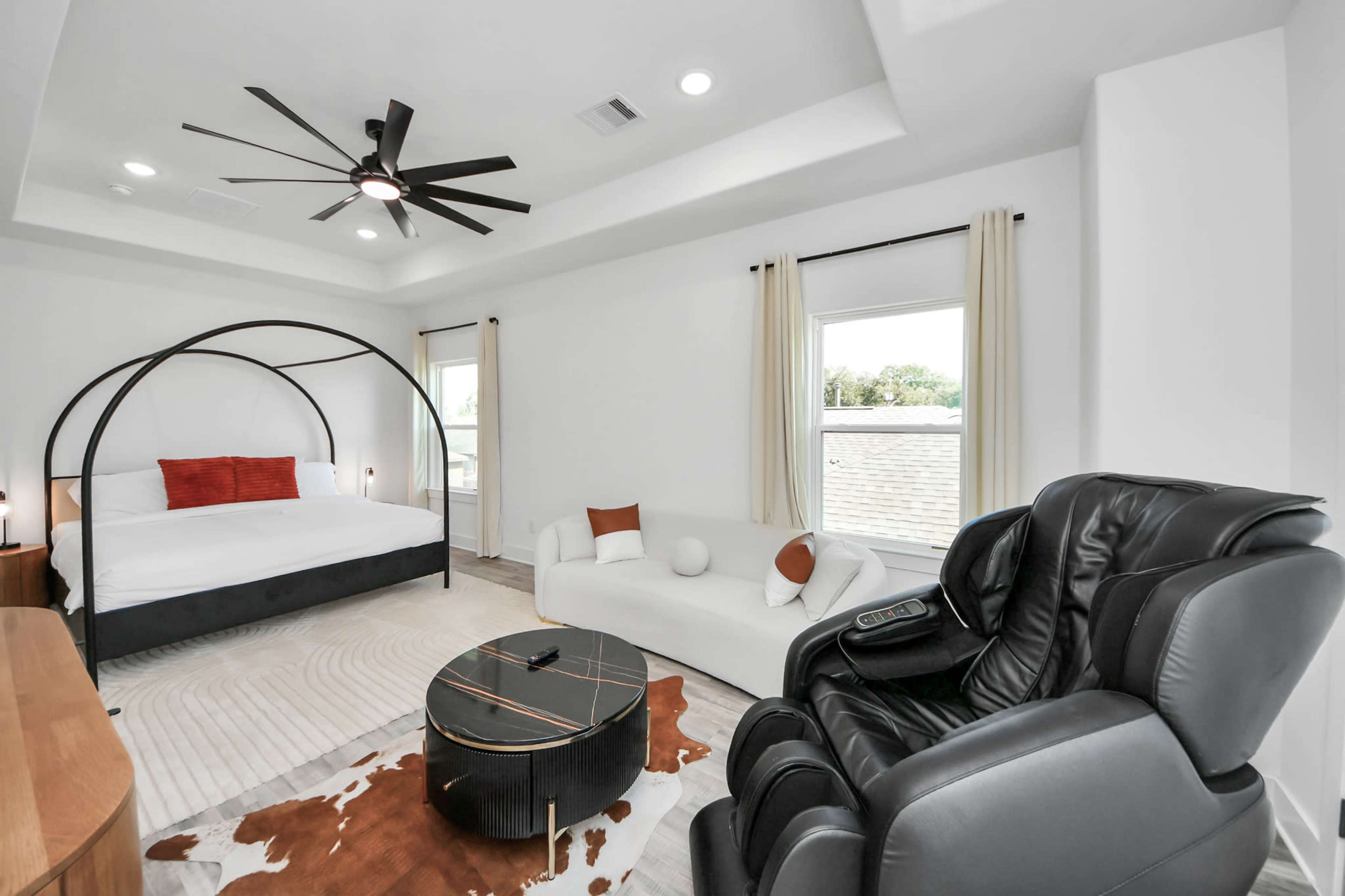 The image shows a modern bedroom featuring a large bed with an arched frame, a sectional sofa, a recliner, and a round coffee table on a patterned rug.