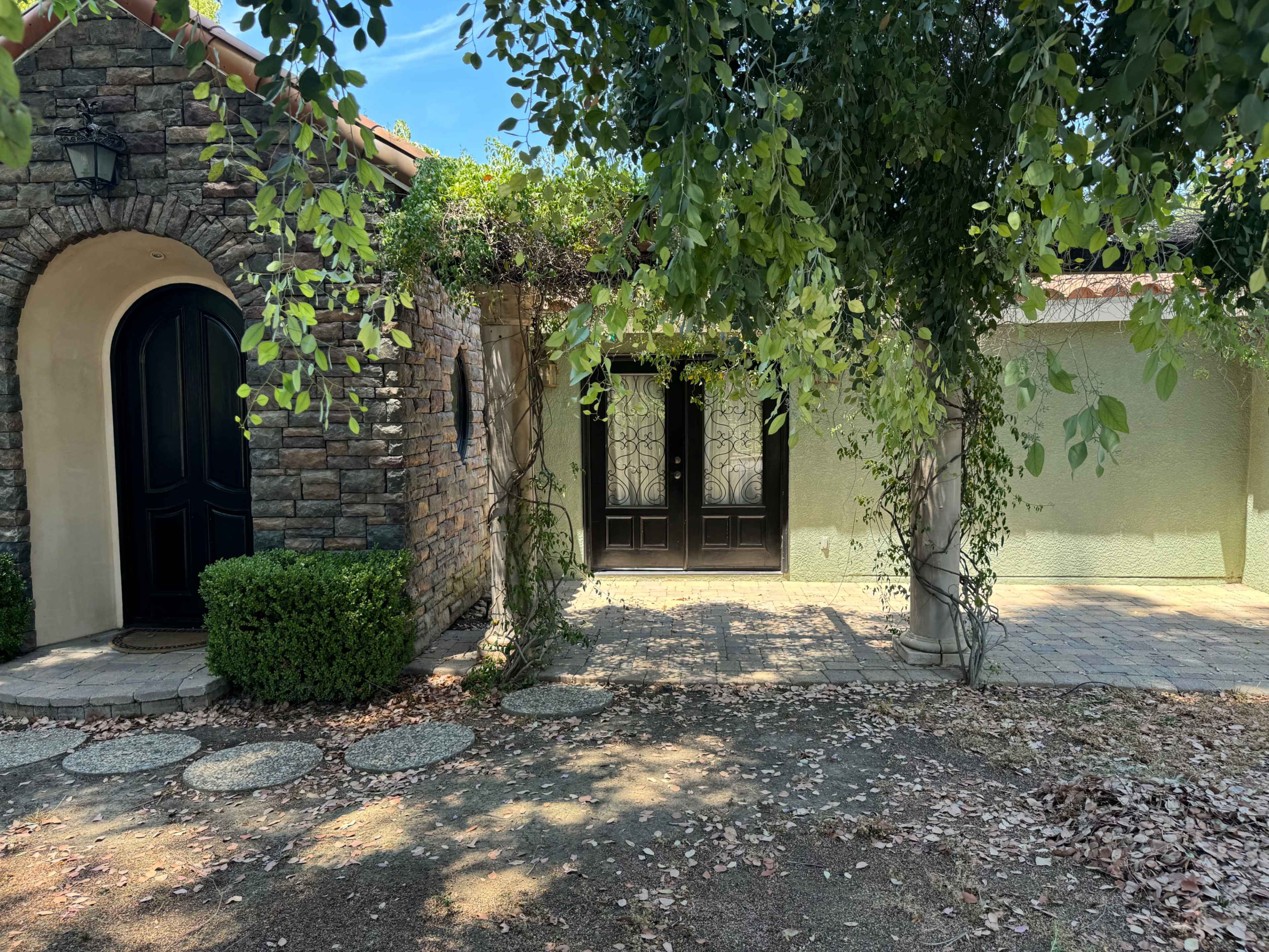 A stone and stucco home entrance with two wooden doors, framed by greenery and a stone pathway.