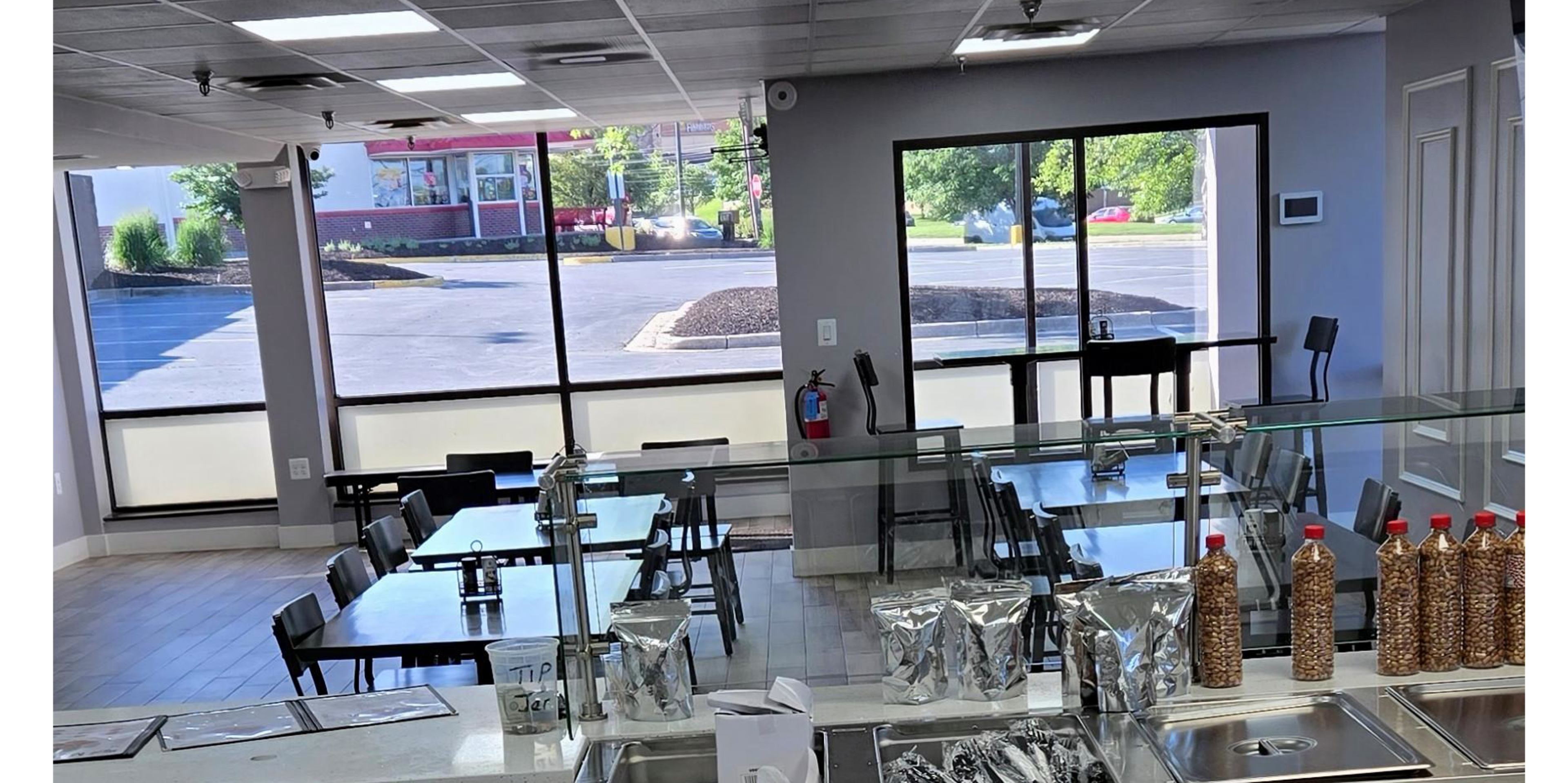 The image shows a bright, modern dining area with tables and chairs set up in front of large windows overlooking an empty parking lot.