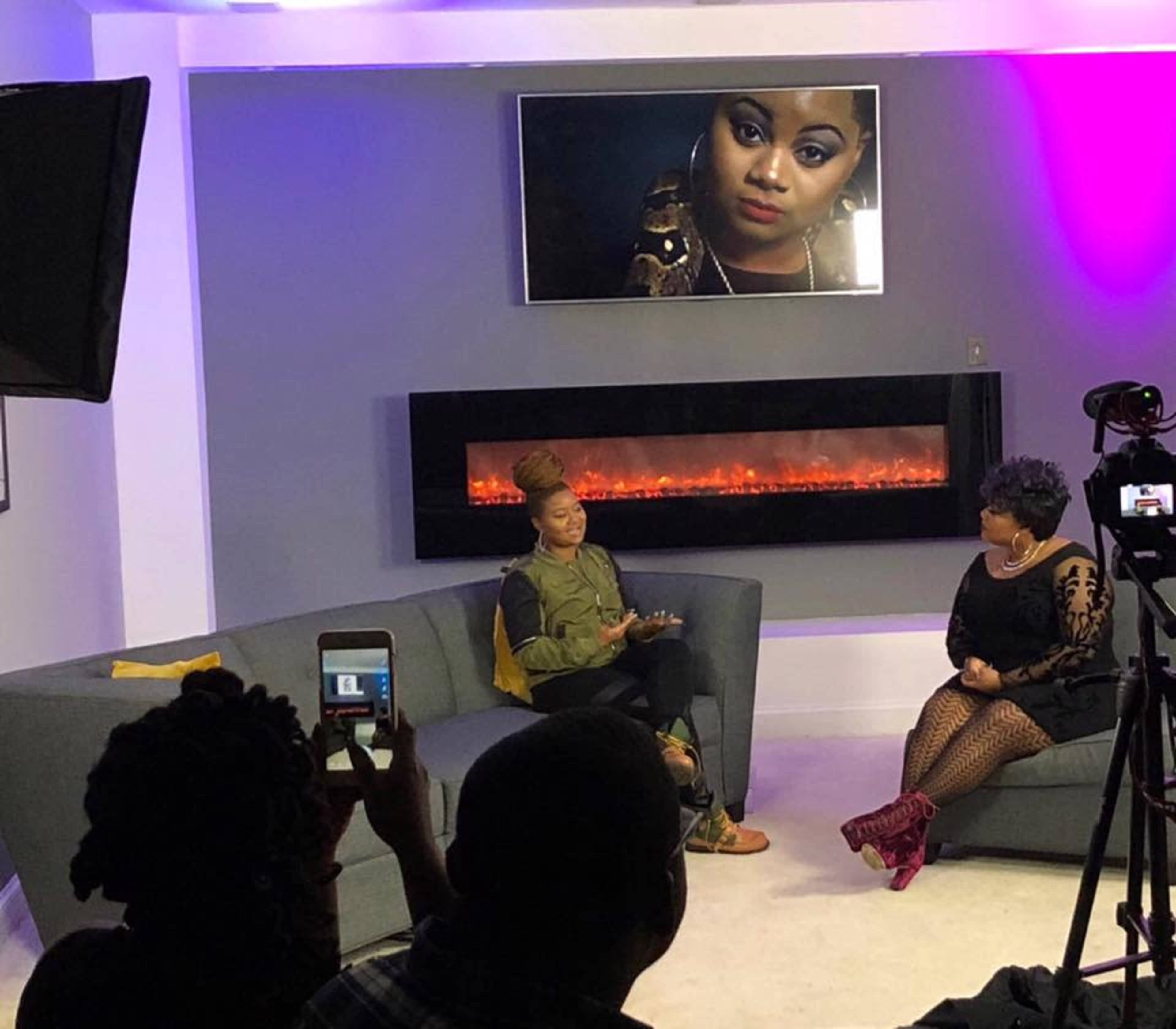 Two women are seated on a gray couch in a modern studio, with a large portrait of a woman displayed on the wall above a glowing electric fireplace.