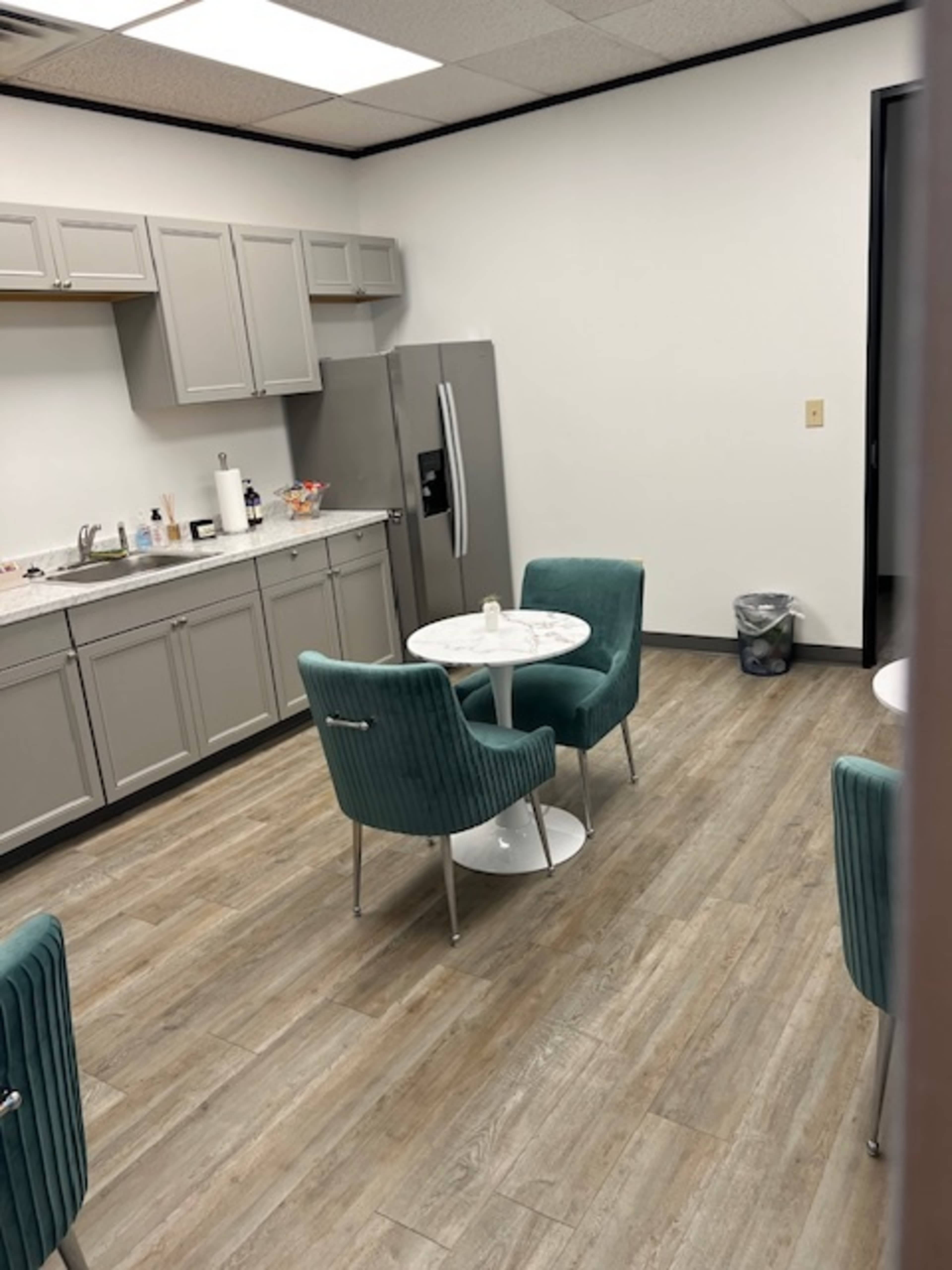 The image shows a break room with a kitchenette featuring gray cabinets, a refrigerator, and a table surrounded by four teal chairs.