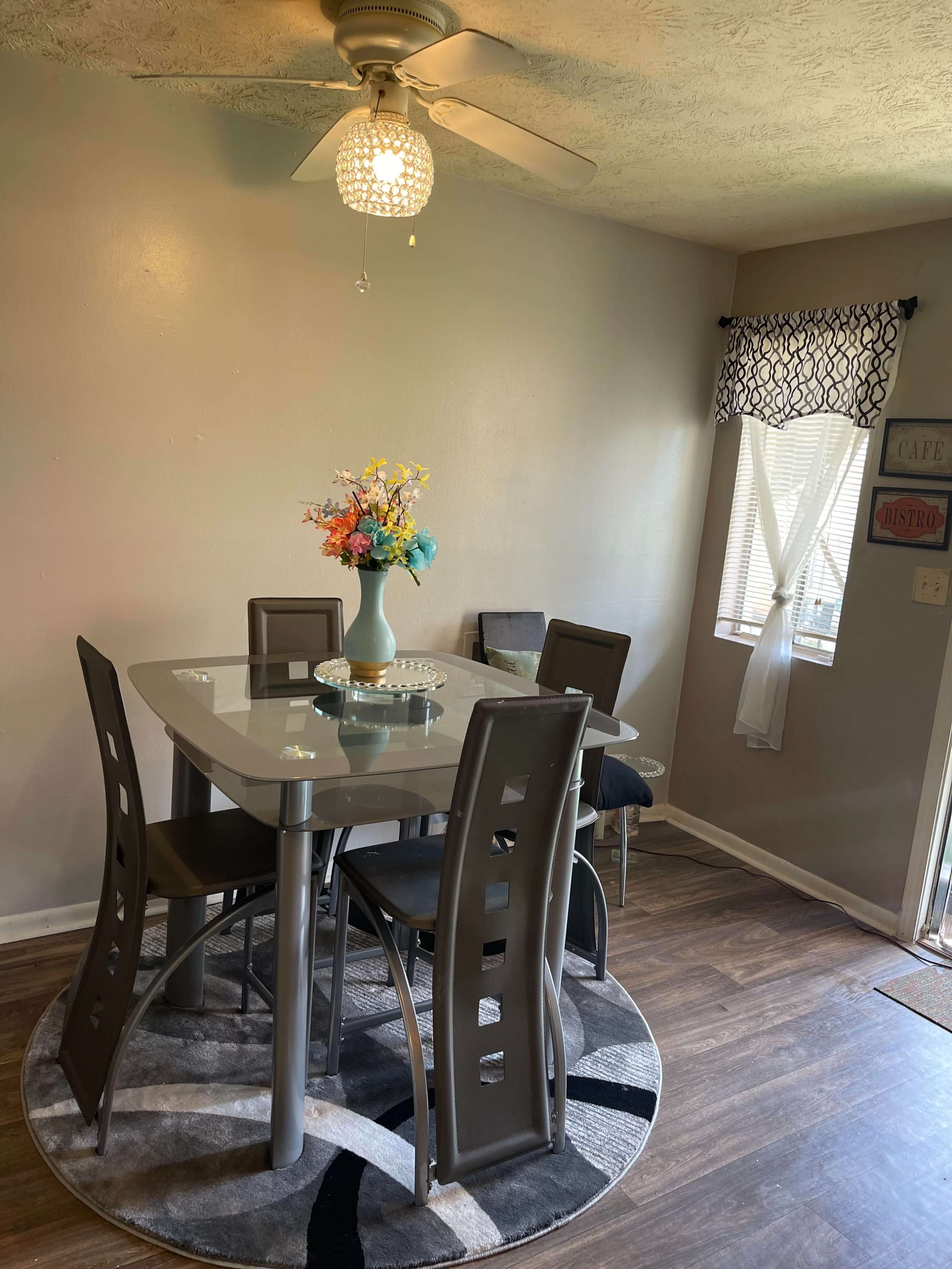 A modern dining area features a glass-top table surrounded by four chairs, a decorative vase with flowers, and a circular rug under the table.
