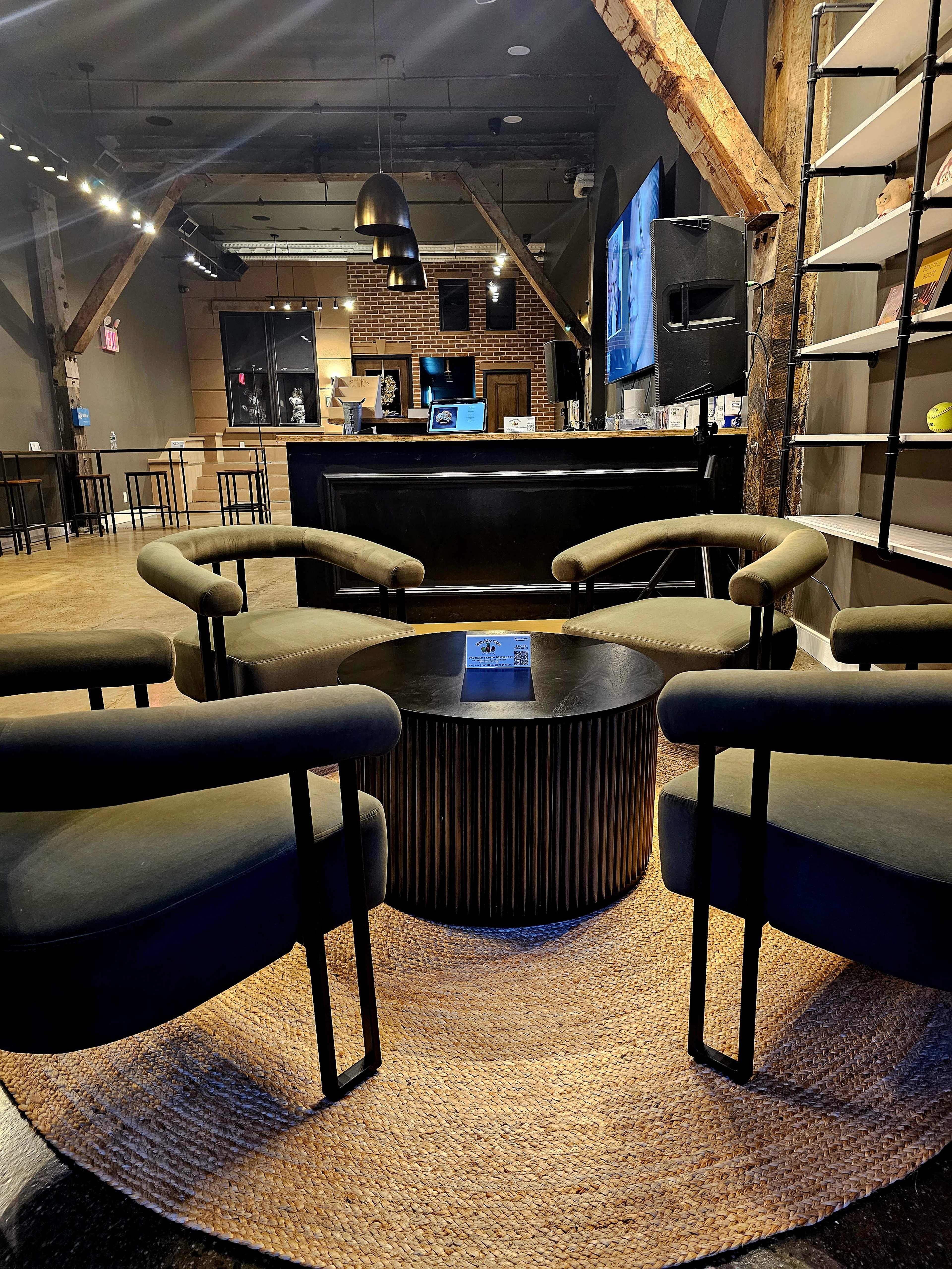 The image shows a modern lounge area with four black chairs surrounding a round wooden coffee table on a textured rug, set against a backdrop of exposed wooden beams and a bar counter.
