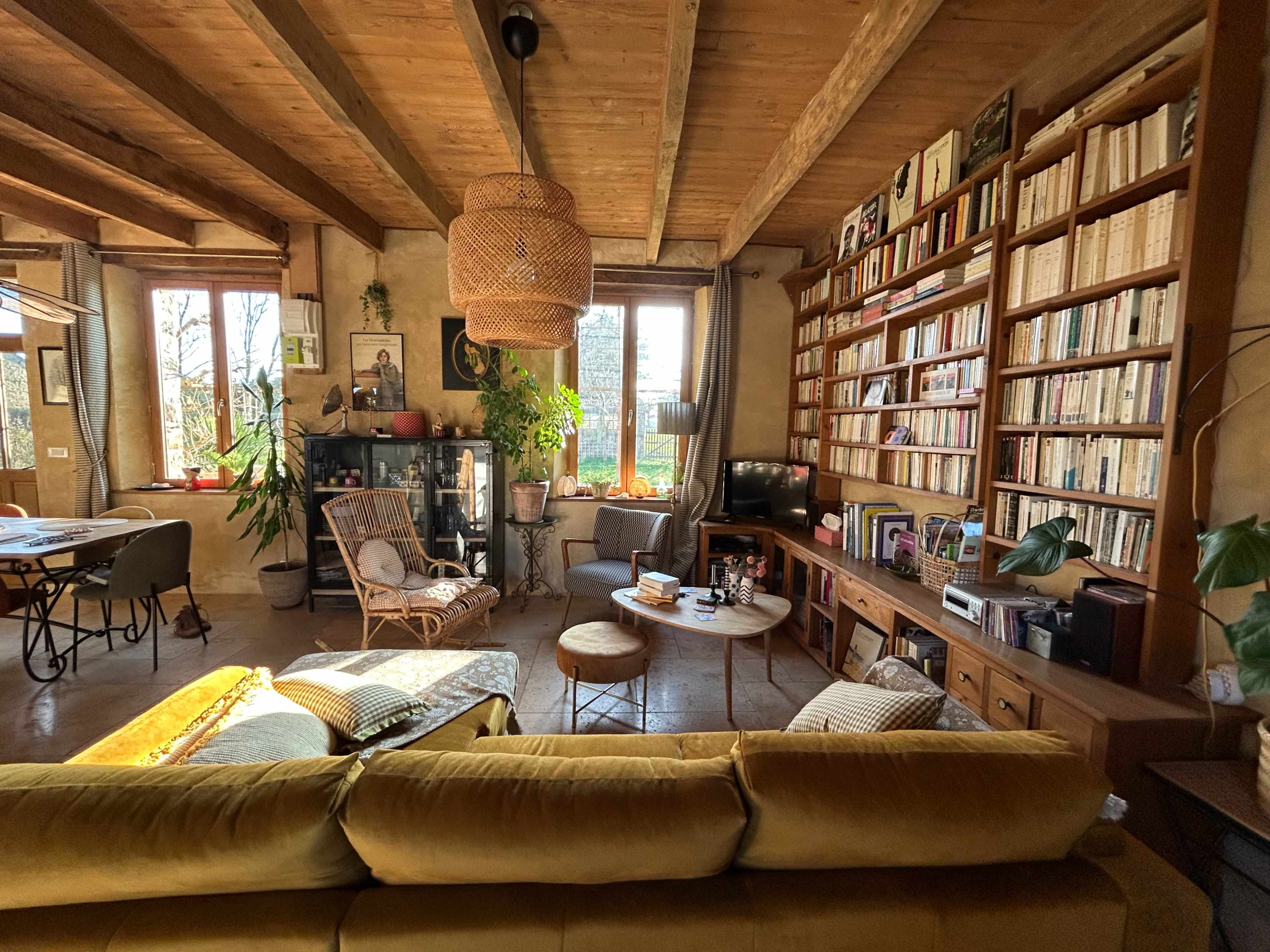 The image shows a cozy living room with wooden beams, a large bookshelf filled with books, and a comfortable yellow sofa facing a small table.