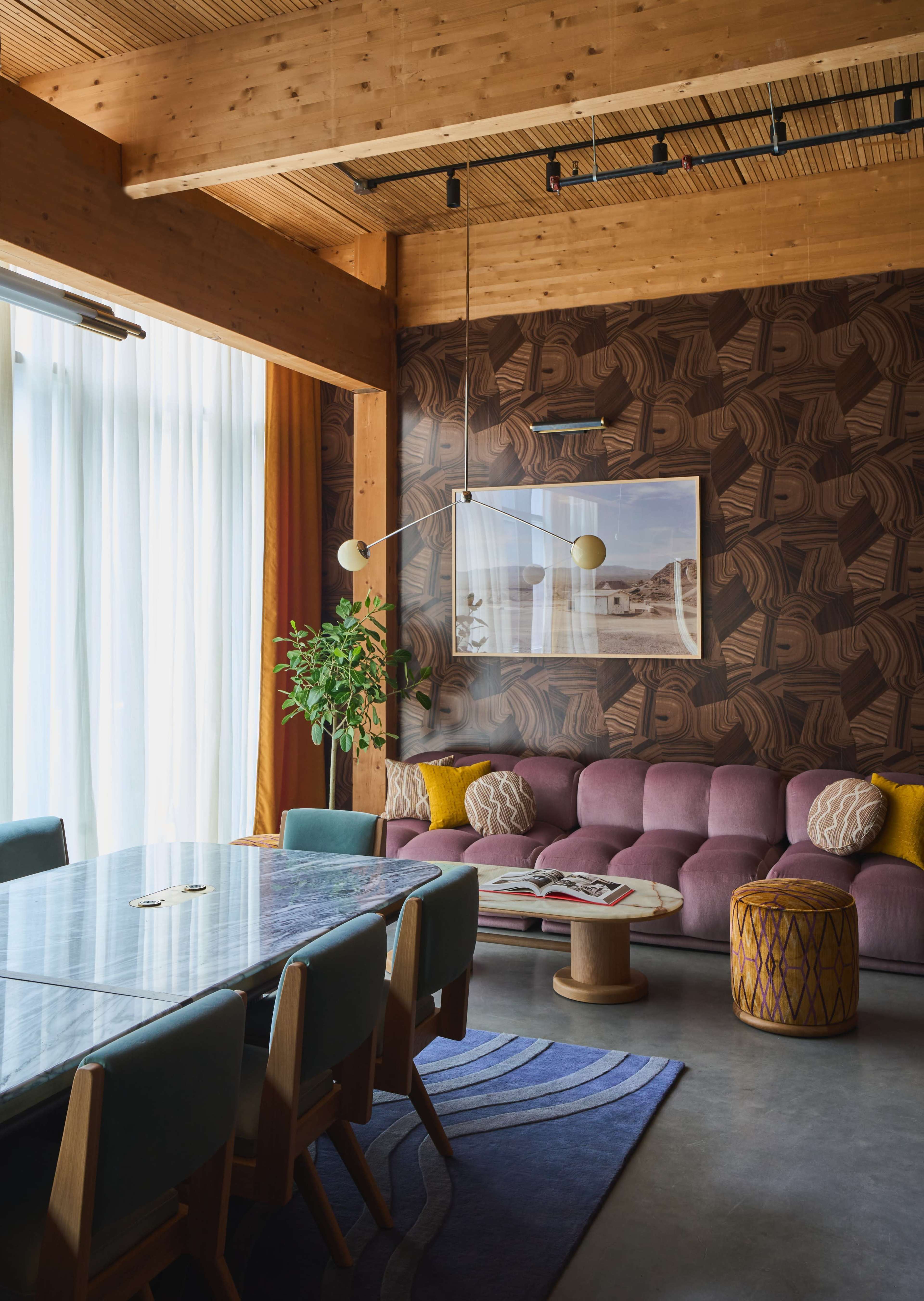 A modern living space with a purple sofa, a wooden dining table surrounded by light blue chairs, large windows, and decorative plants.