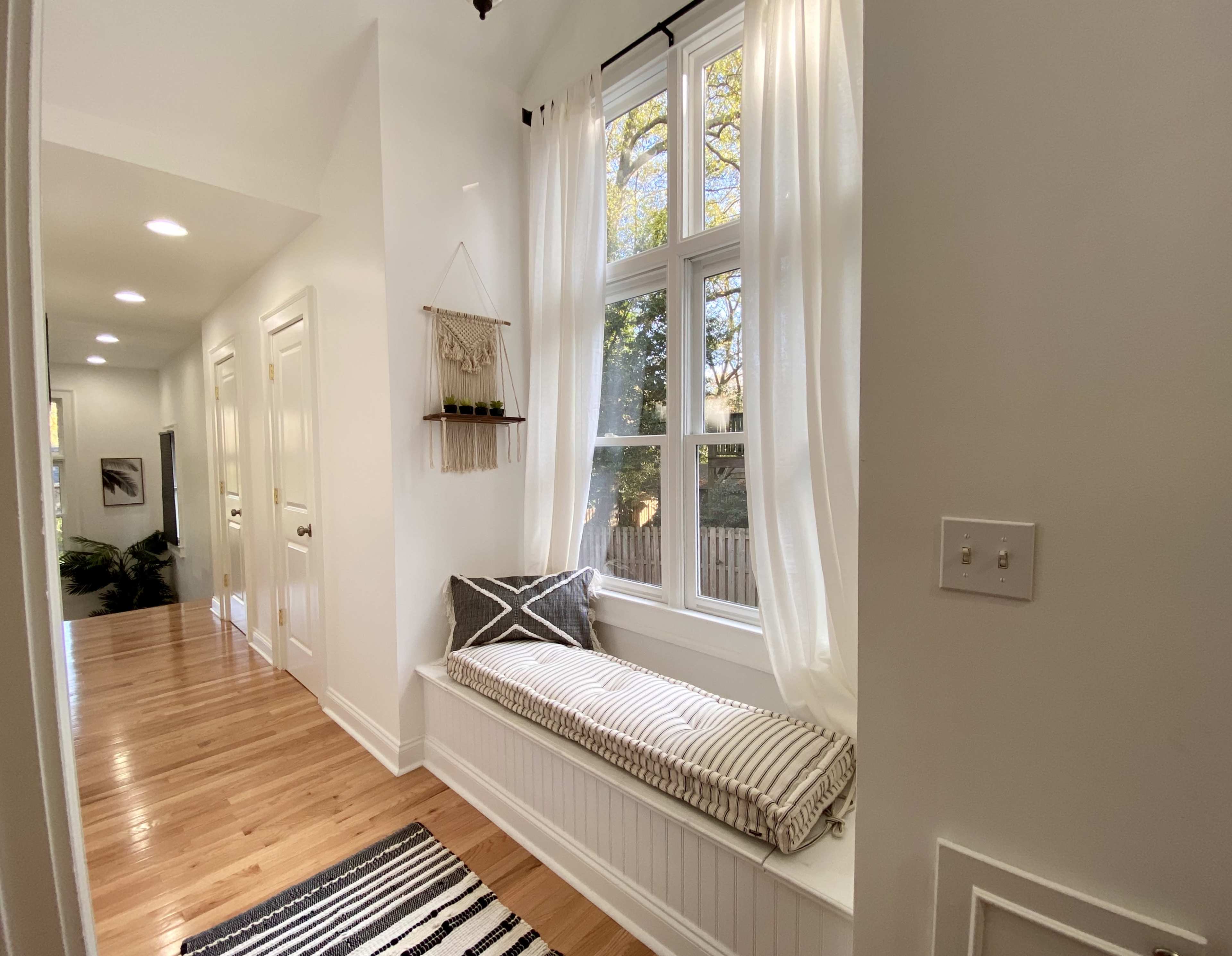 A bright hallway features a window seat with a striped cushion, framed by white curtains and adjacent to wooden flooring and a decorative wall hanging.