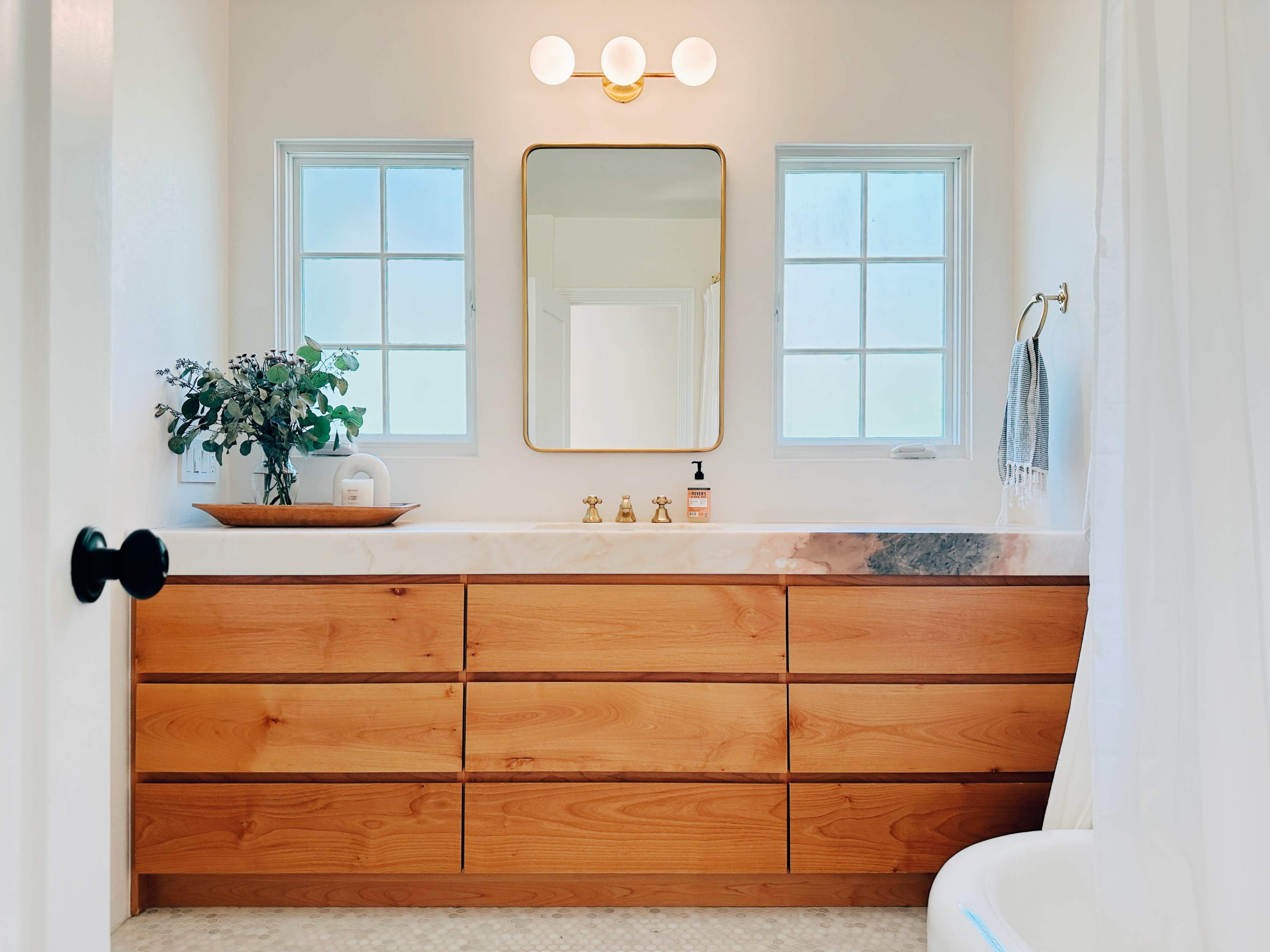 A modern bathroom featuring a wooden vanity with multiple drawers, a round mirror, two windows, and a light fixture above.
