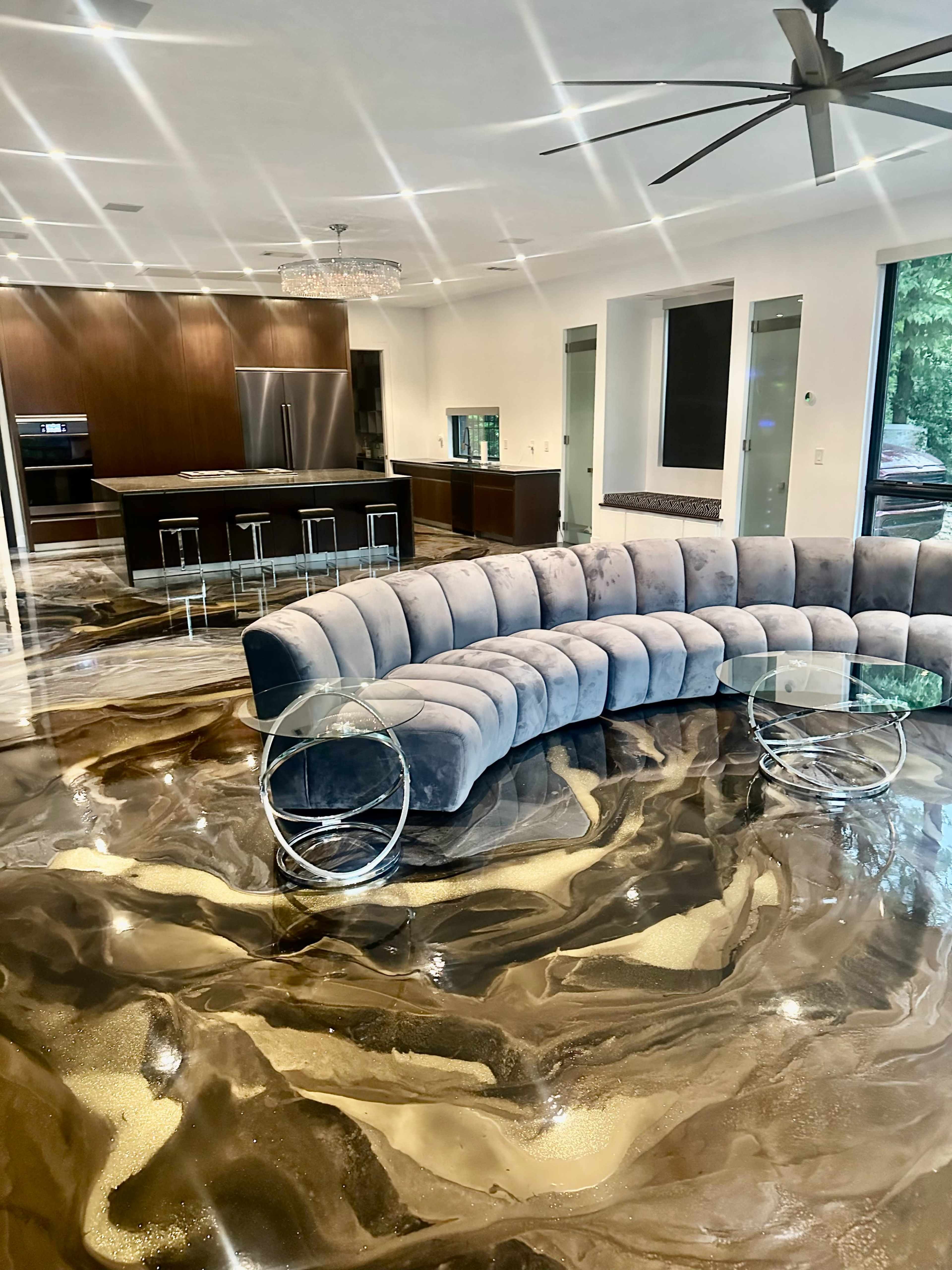 The image shows a modern living area featuring a curved gray couch, a glass coffee table, and a kitchen in the background with dark cabinetry and stainless steel appliances, highlighted by polished, marble-like flooring.