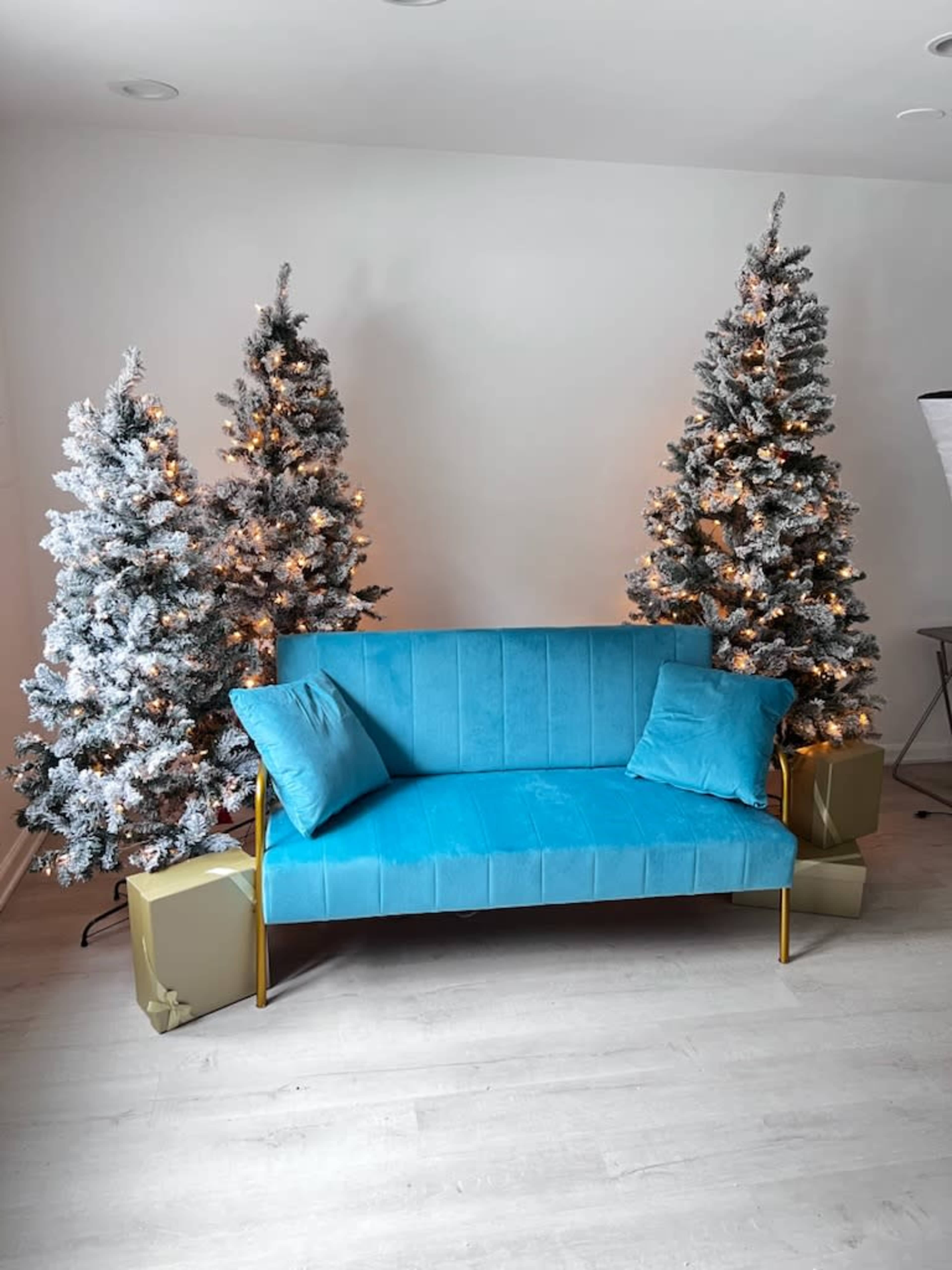 A bright blue couch is positioned between two snowy Christmas trees, each adorned with warm lights, and the floor is decorated with wrapped gifts.