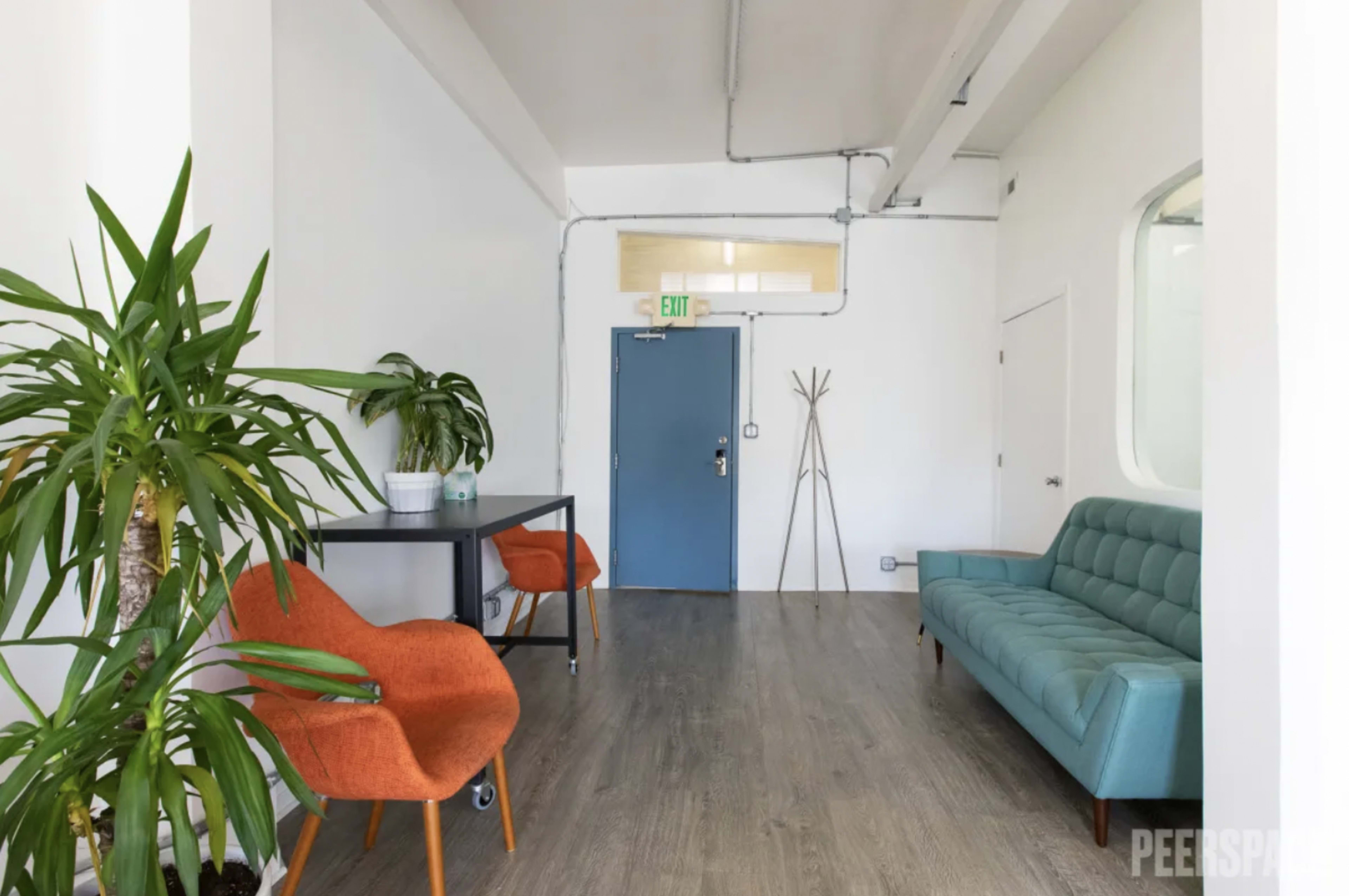 The image shows a minimalist interior space with a teal couch, orange chairs, a small table with a plant, and a blue door.