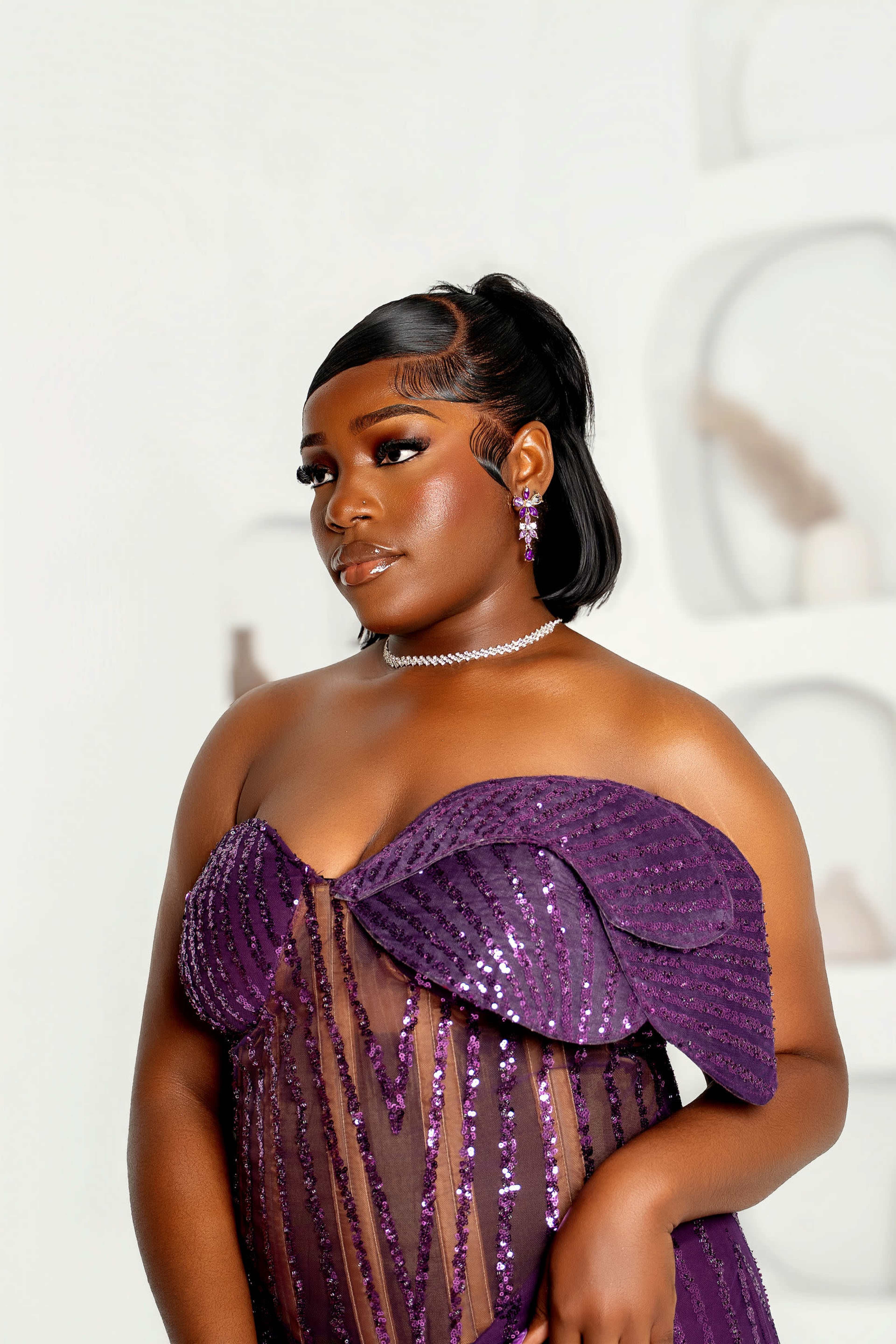 A woman wearing a sparkling purple gown with a sheer overlay poses in front of a minimalist backdrop.